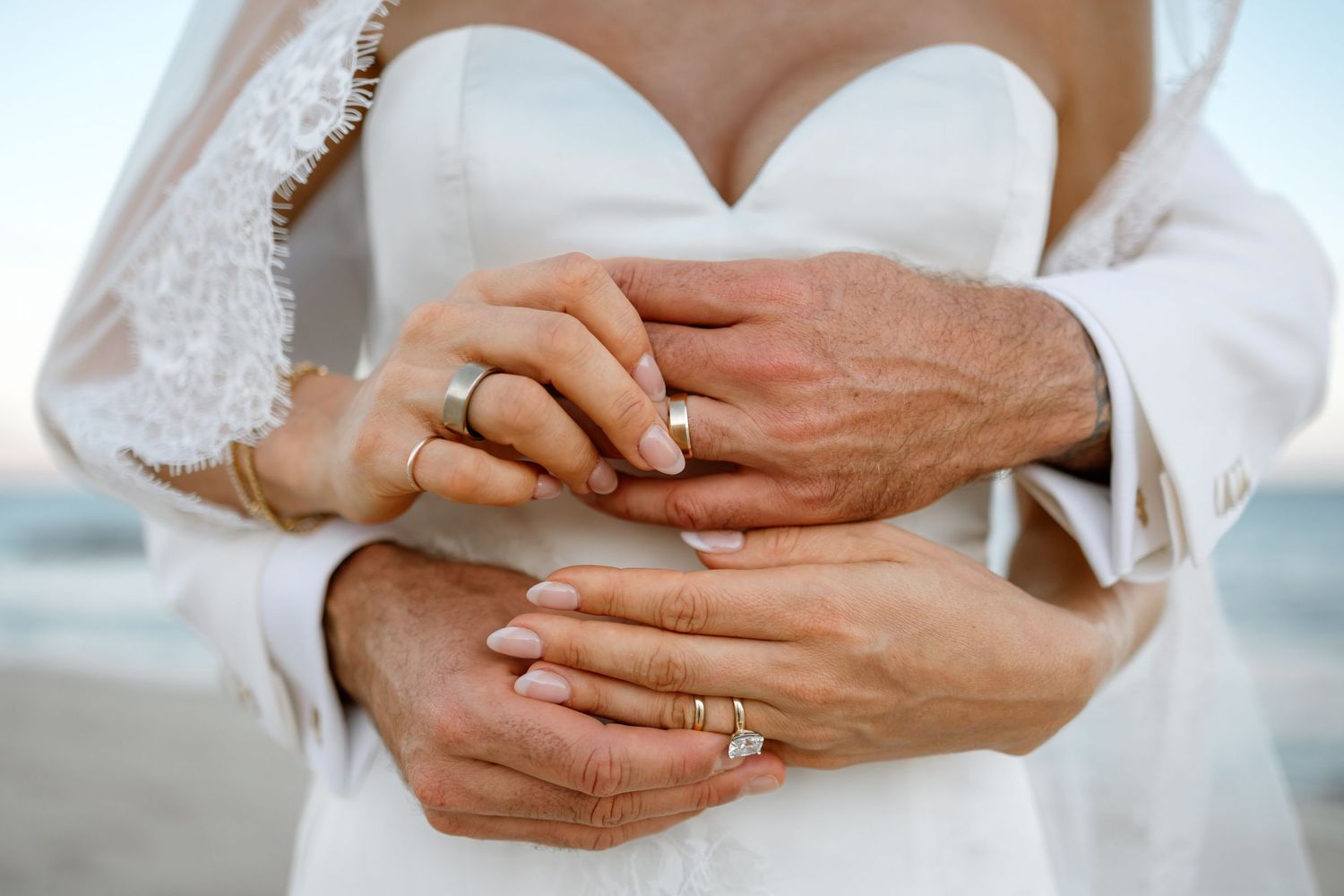 Bride and groom showing wedding rings at beach ceremony, hands embracing in white wedding dress.