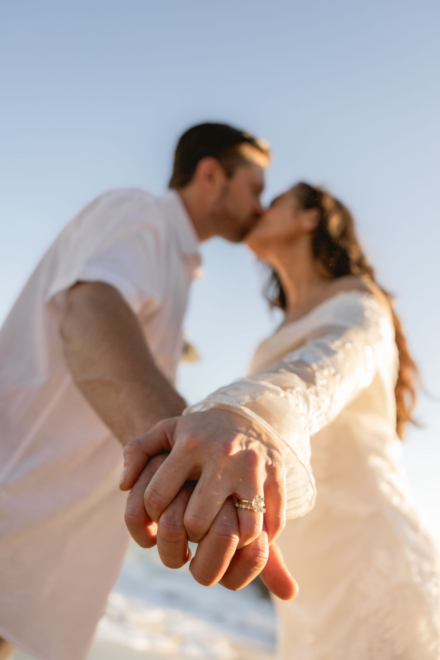 A close-up of a couple kissing on the beach with an engagement ring visible.