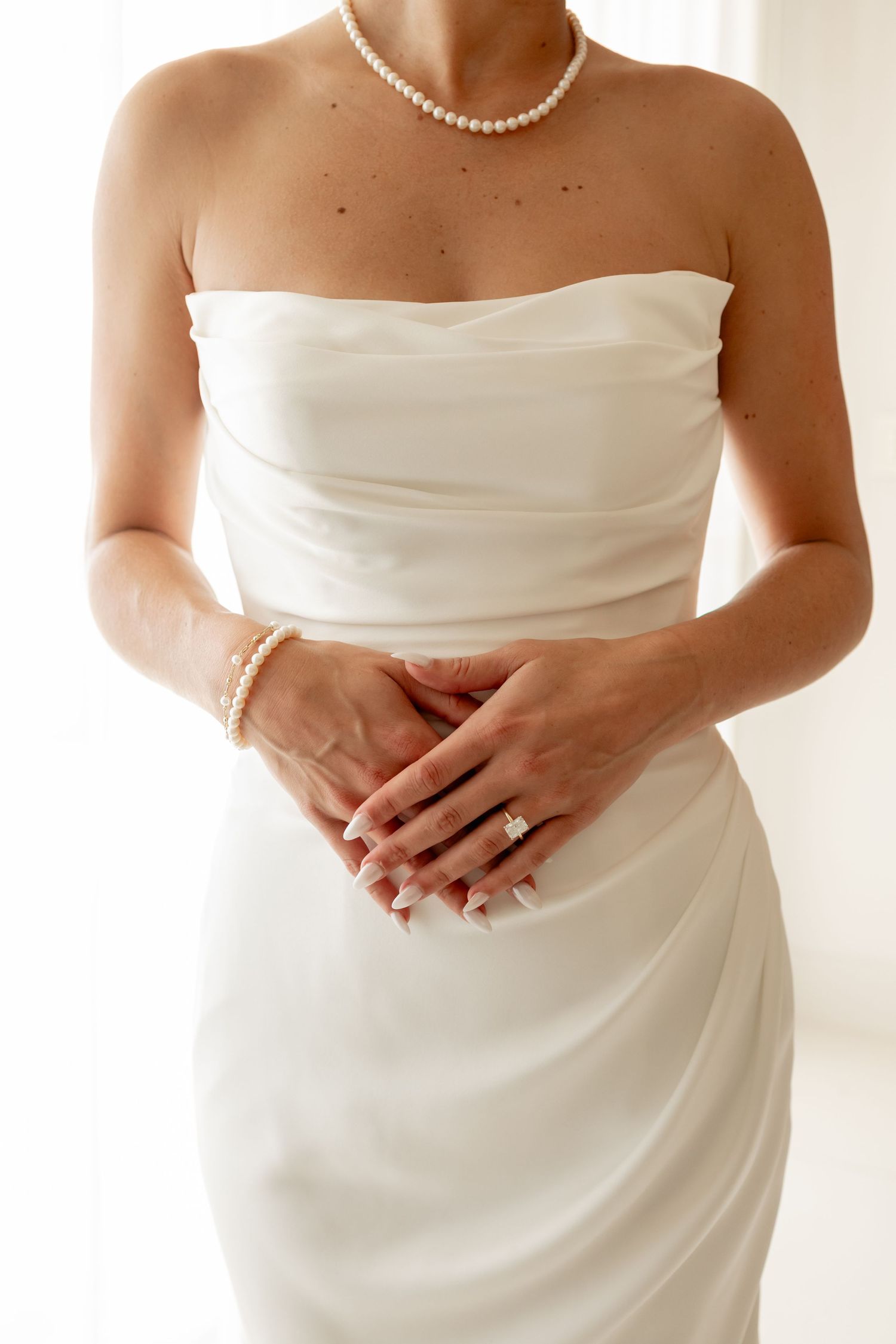 Bride in strapless white satin gown with pearl necklace, bracelet, and diamond engagement ring, hands clasped.