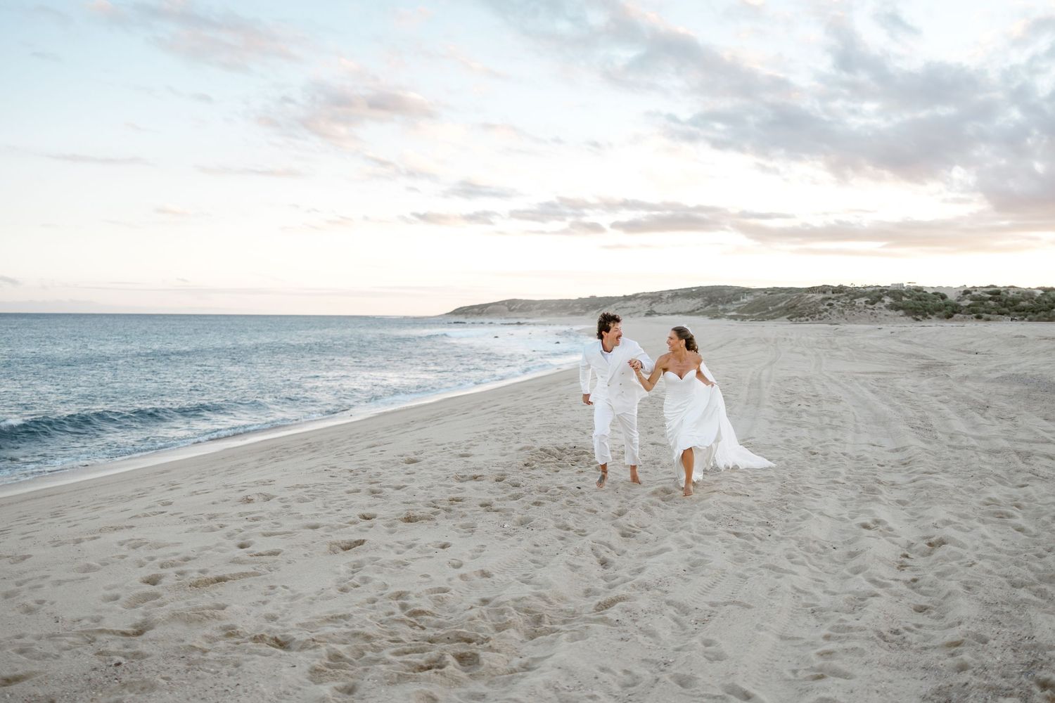 A couple in white outfits walking together on a serene beach at sunset while holding hands.