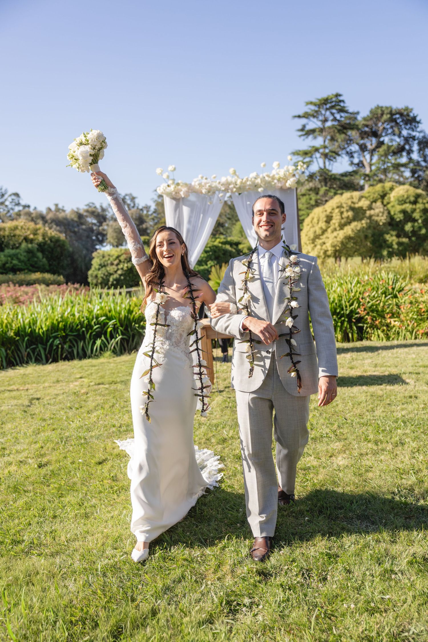 Newlyweds celebrating on sunny lawn with confetti and white floral decorations.