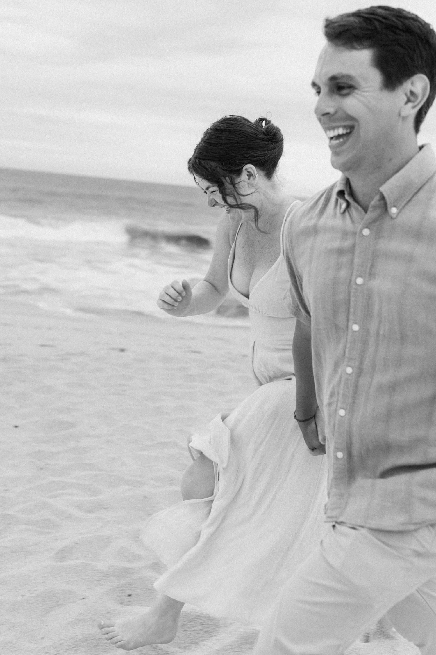 A couple in white clothing walks joyfully along a beach shoreline in black and white photography.