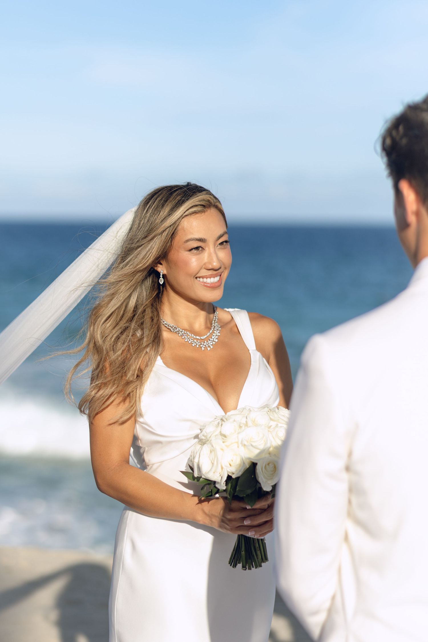 Bride in white dress holds bouquet during beachside wedding ceremony.