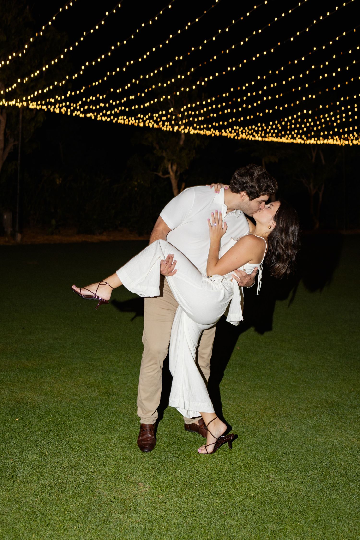 A couple in white and beige dance together under twinkling string lights at an outdoor evening reception.