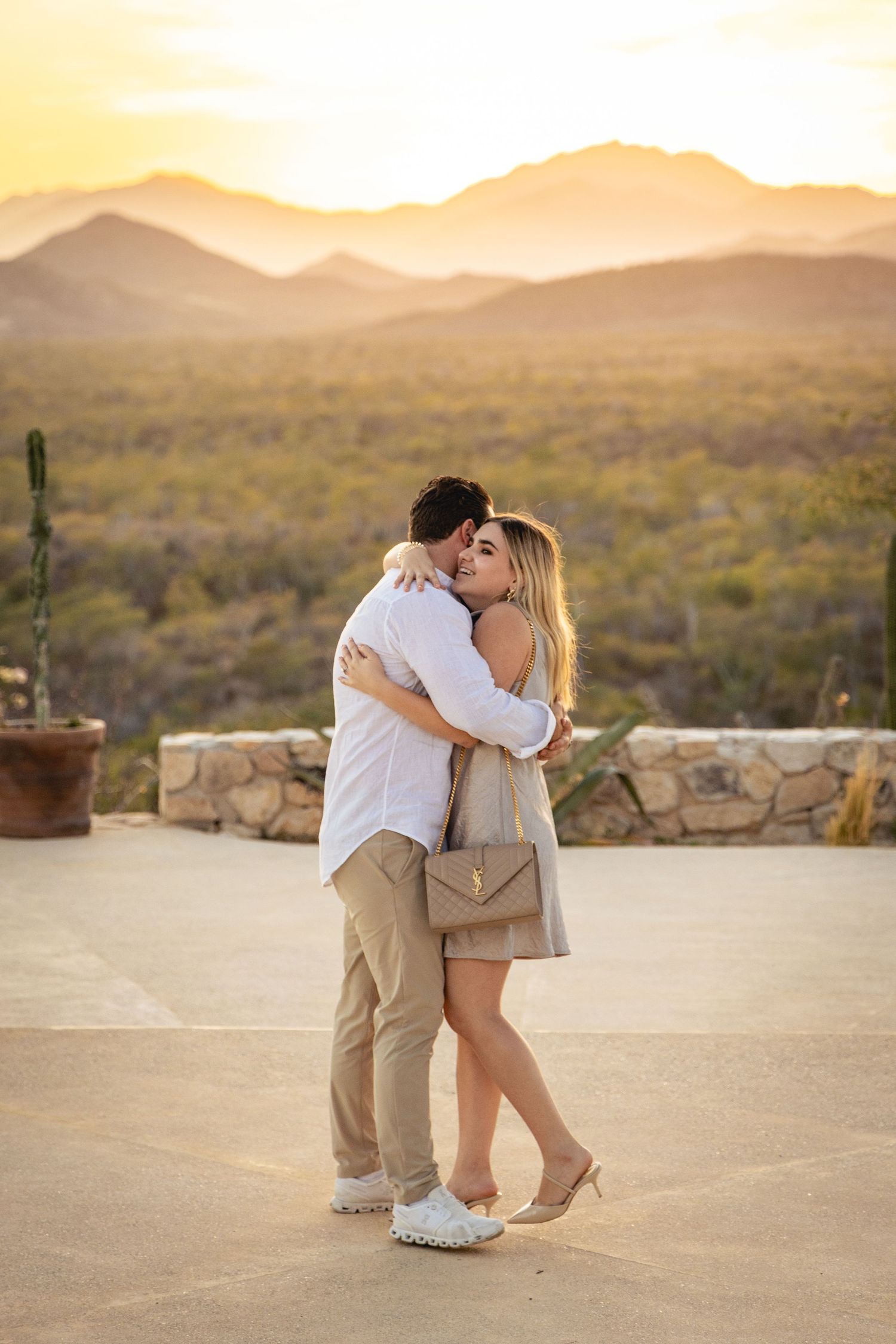 A couple embraces in a romantic moment at sunset with desert mountains and cacti in the background.
