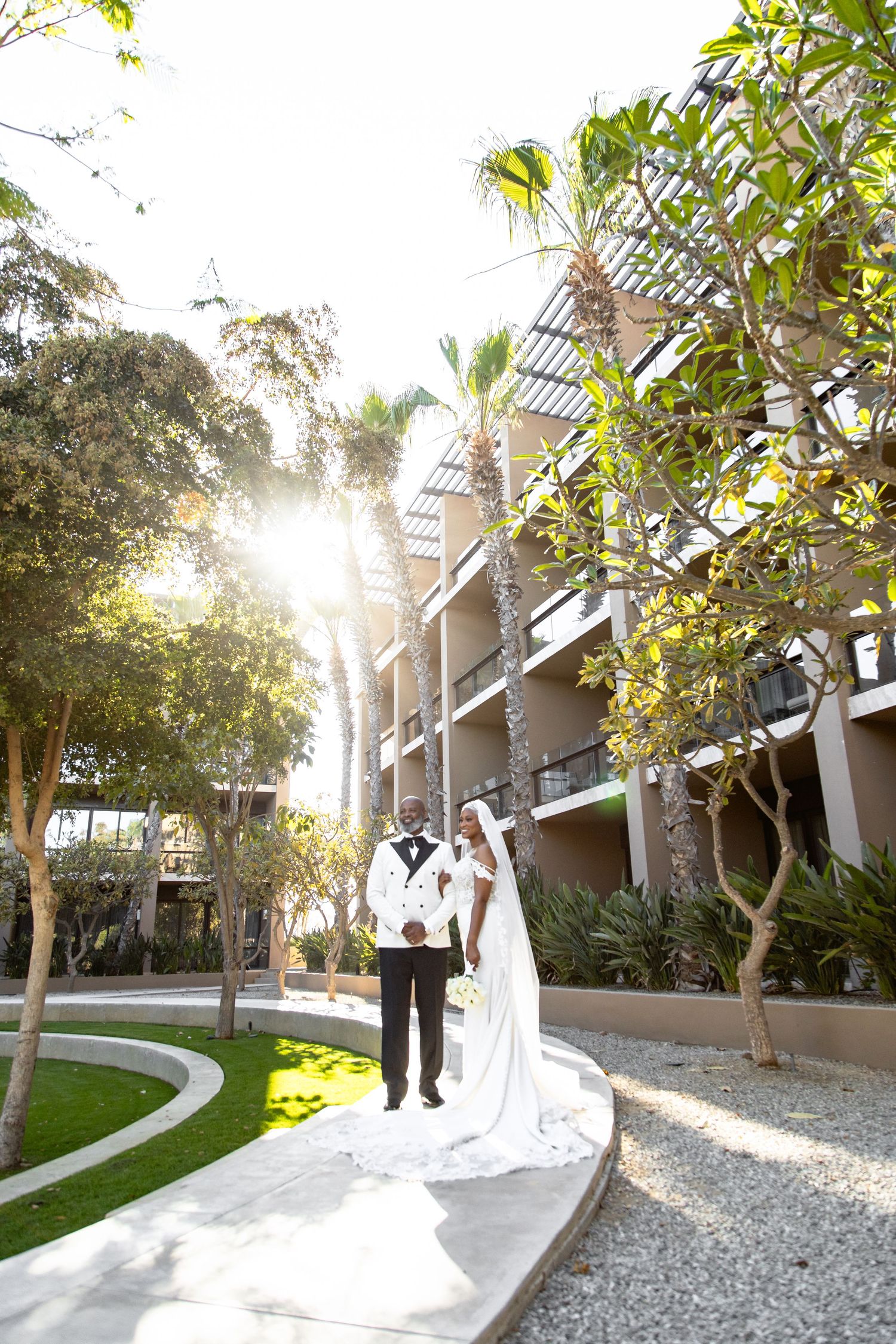 Wedding couple walking through garden pathway with sunlight streaming through trees.
