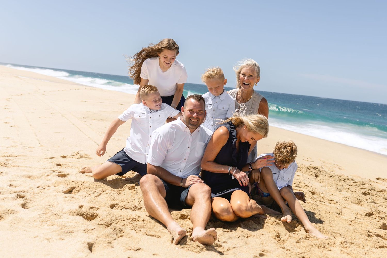 A joyful group sits together on a sunny beach with waves and blue ocean in the background.