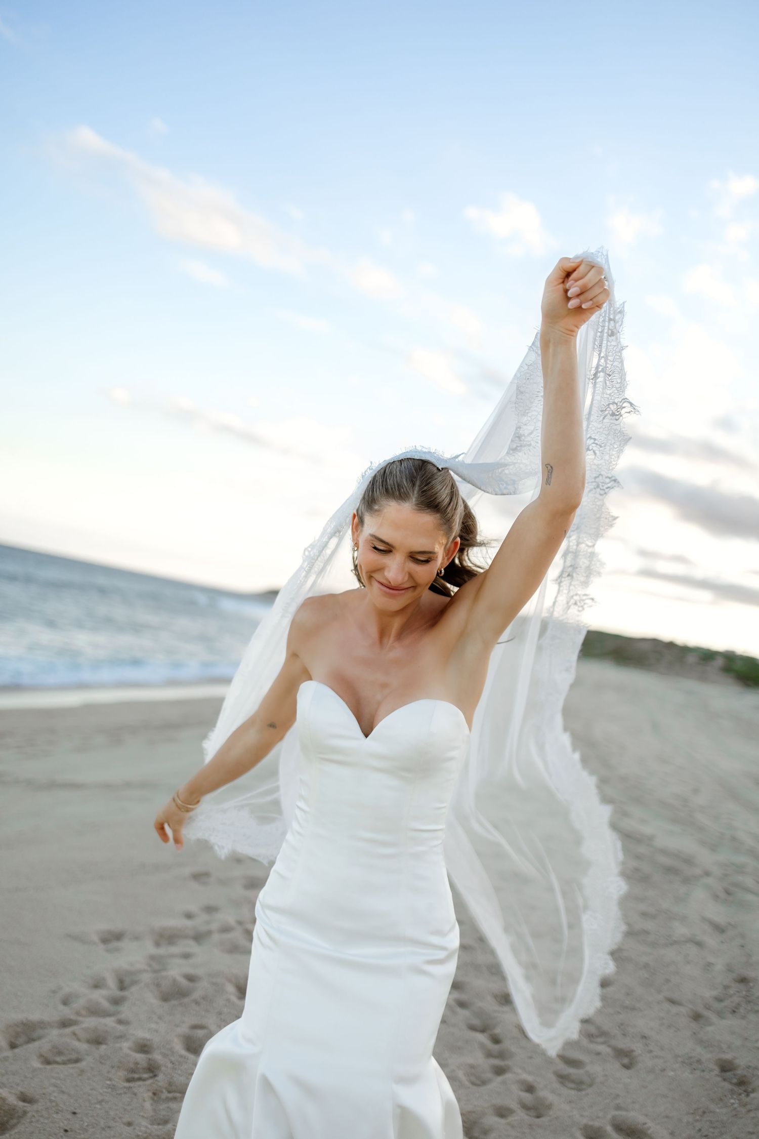 Bride in flowing white dress and veil dancing joyfully on beach.