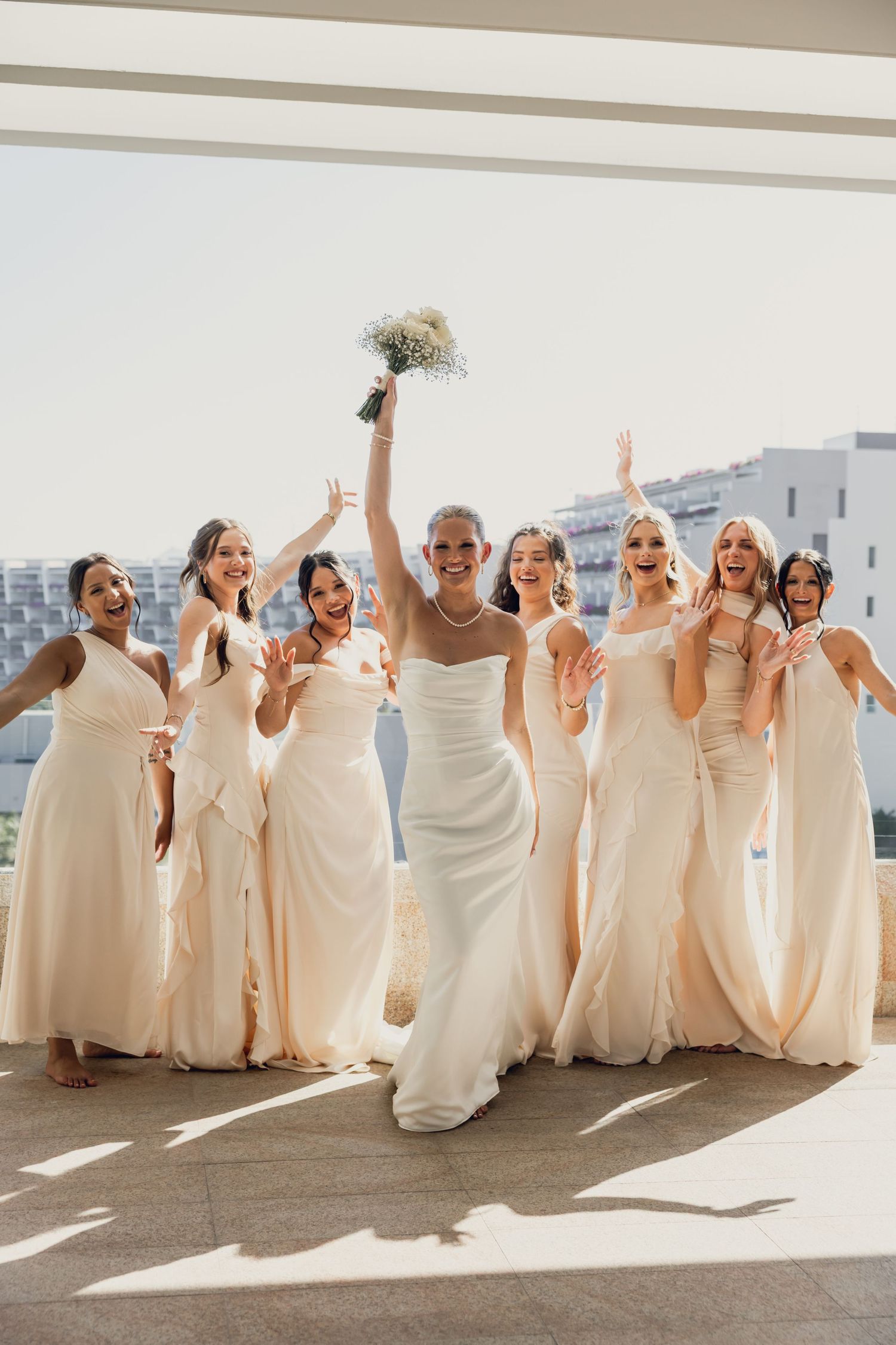 Bride in white gown holds bouquet aloft with six bridesmaids in cream dresses celebrating on a sunny terrace.