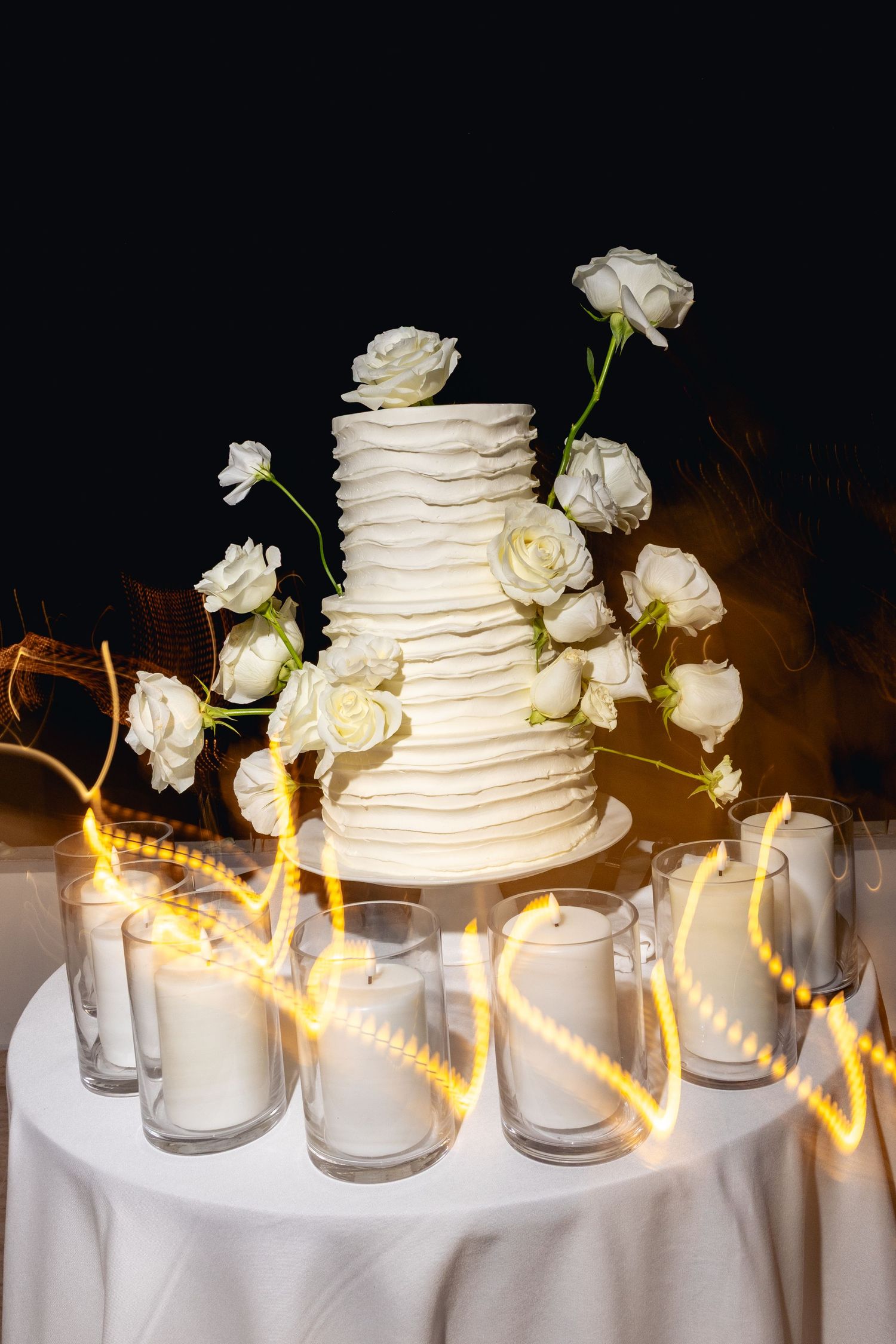 Elegant three-tier white wedding cake adorned with roses, surrounded by glowing candles on a white tablecloth.