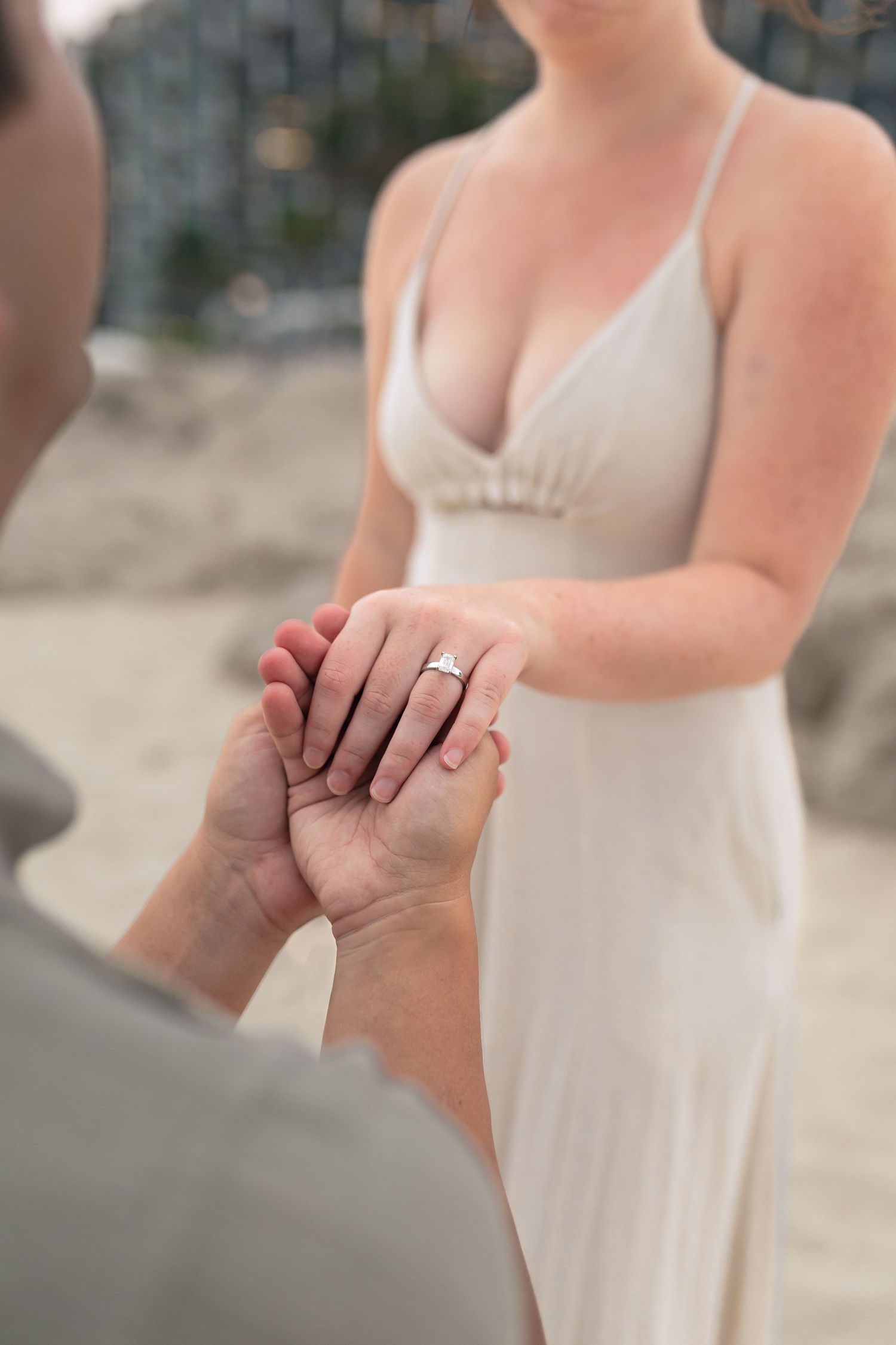 A close-up shot of hands clasped together showing an engagement ring, with a person wearing a white dress in the background.