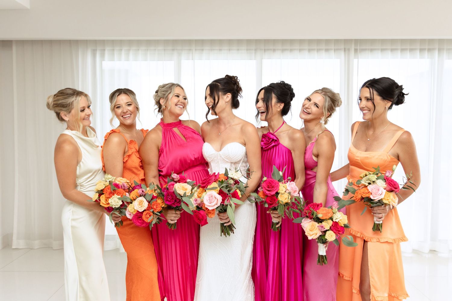 Bridal party in vibrant orange and pink dresses holding colorful floral bouquets.