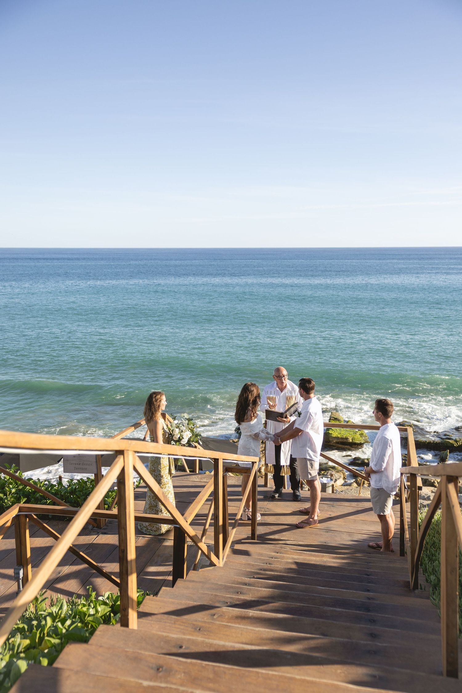 A scenic oceanfront wedding setup with wooden deck railing and guests in white attire.