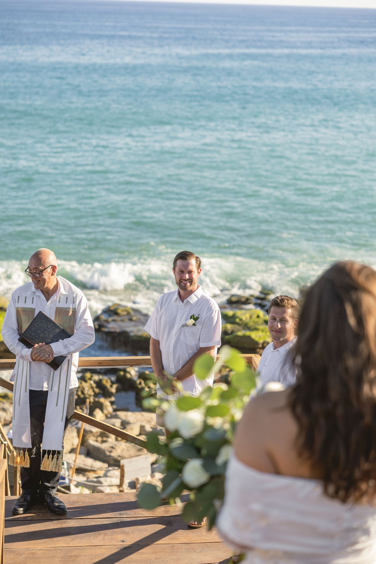 A beachside wedding ceremony unfolds on a wooden deck with ocean waves in the background.