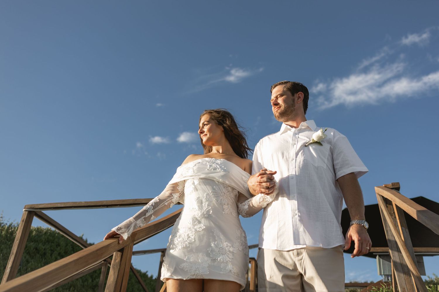 Couple in white outfits descending wooden steps against a blue sky background.