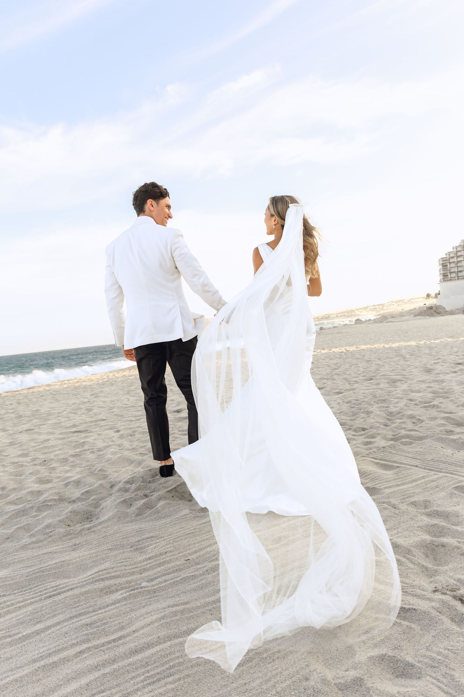 Bride and groom walking playfully along beach shore with flowing white dress and veil catching the wind.