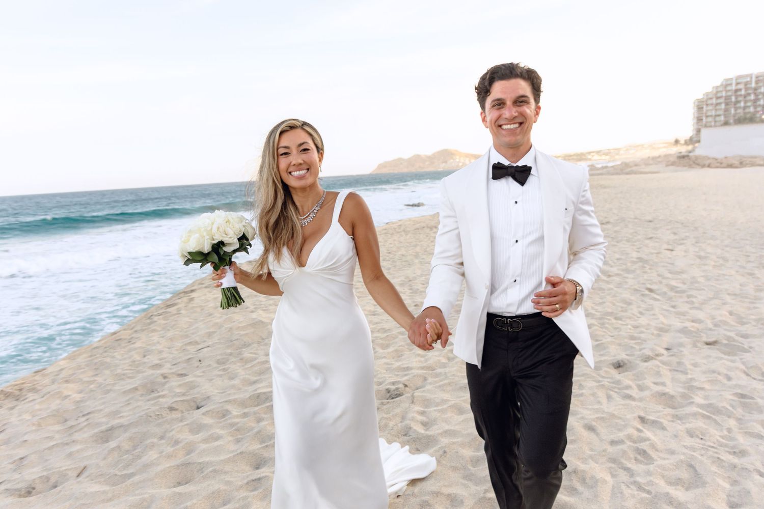 Newlyweds walking hand in hand on beach with bride holding white bouquet.