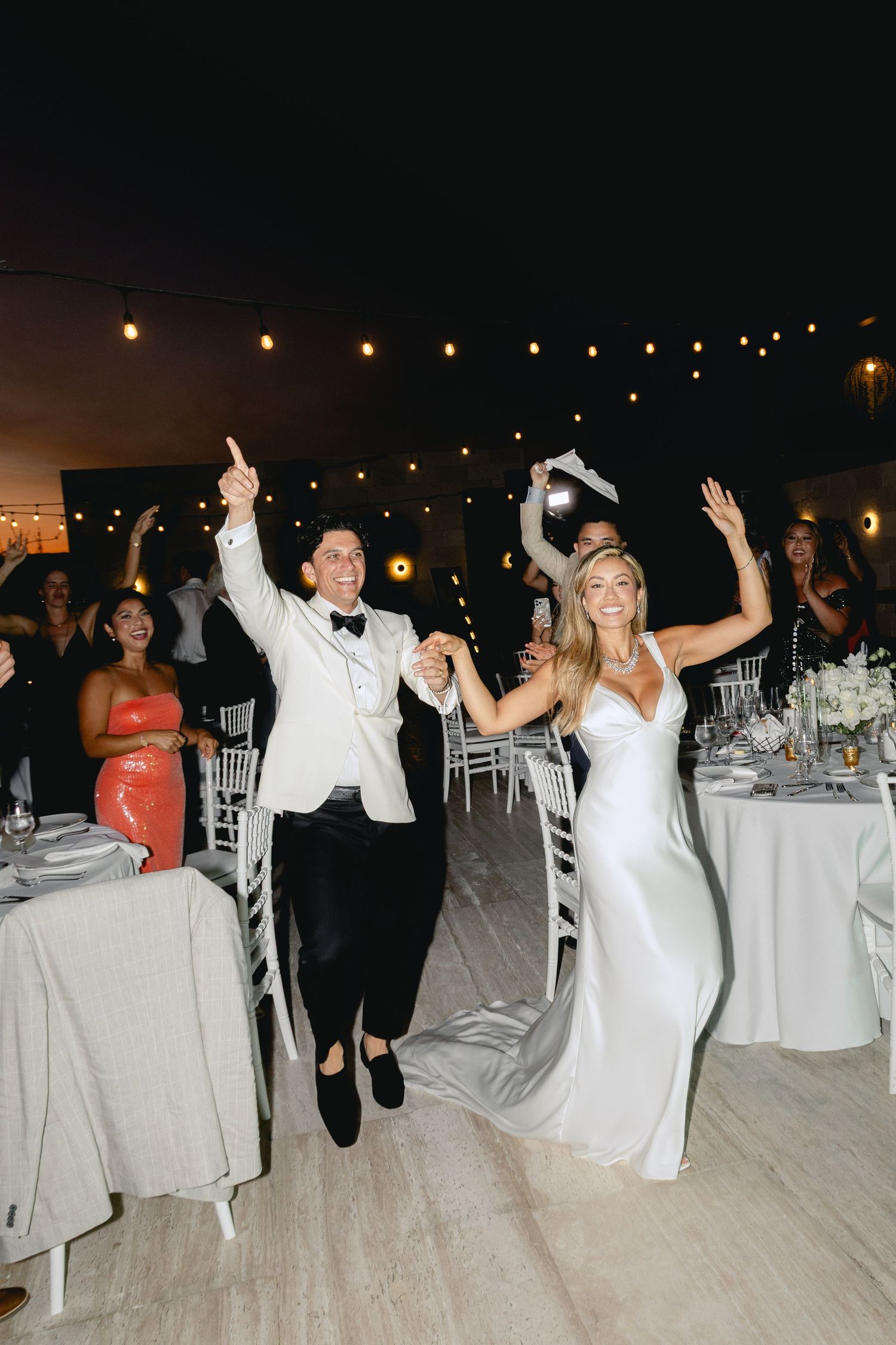 Wedding guests dancing and celebrating at an outdoor reception with white chairs and table settings.