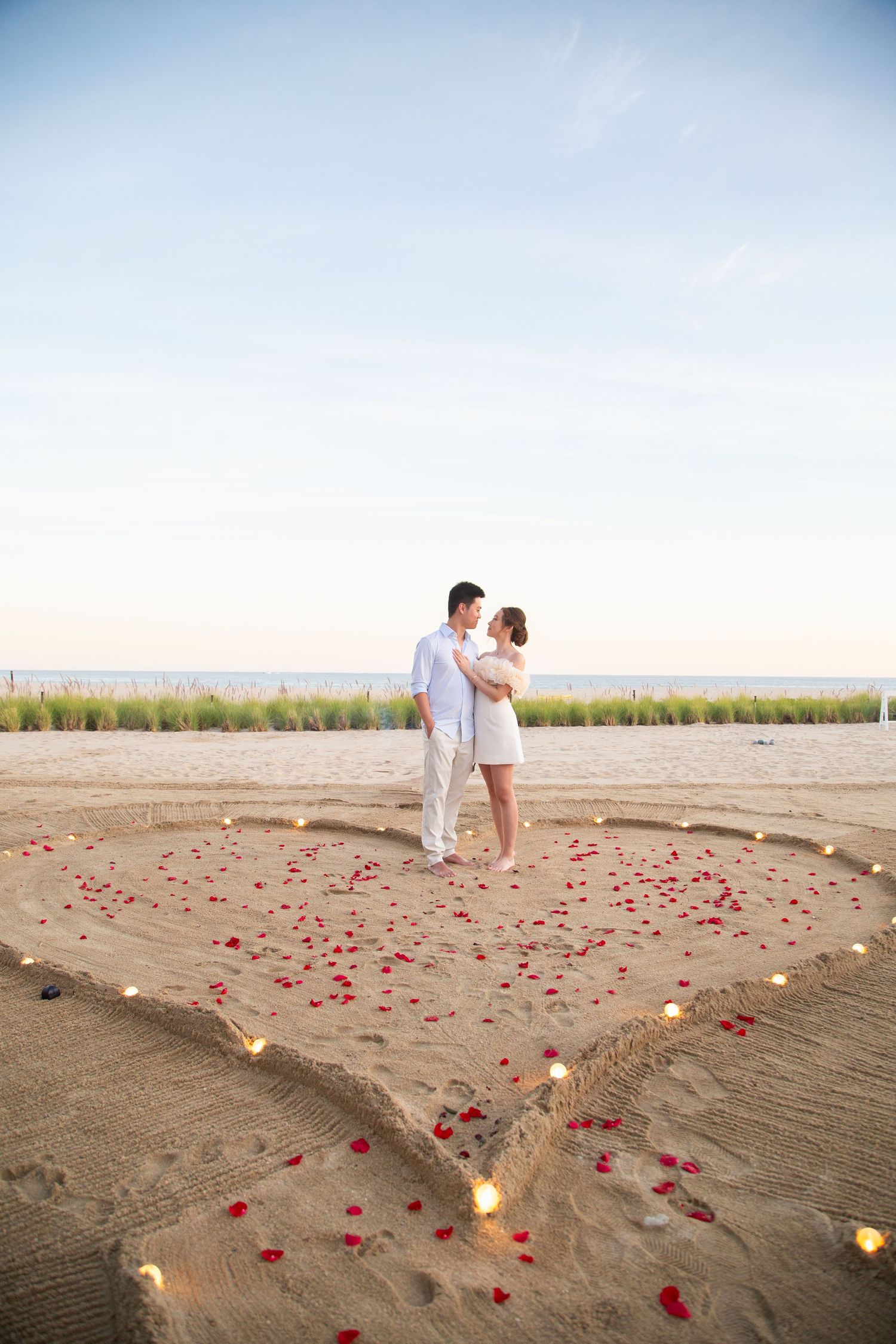 A romantic beach scene with rose petals and candles arranged in a heart shape leading to a couple at sunset.