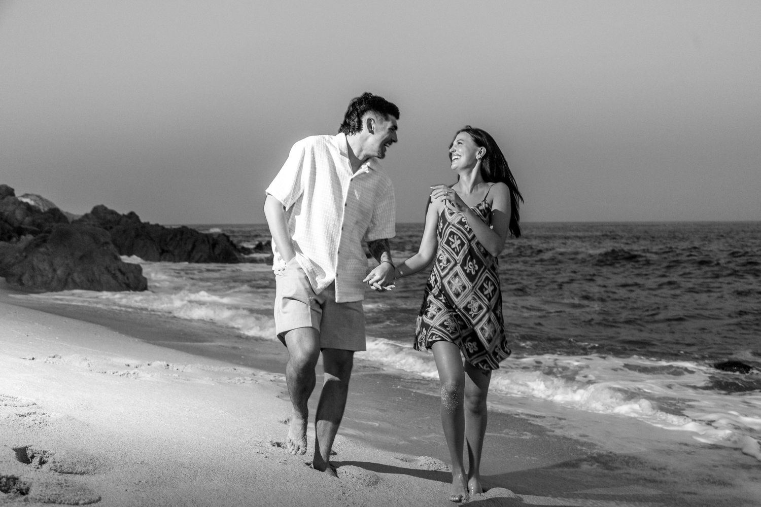 A couple walks along a scenic beach shoreline during sunset, holding hands and enjoying the ocean view.