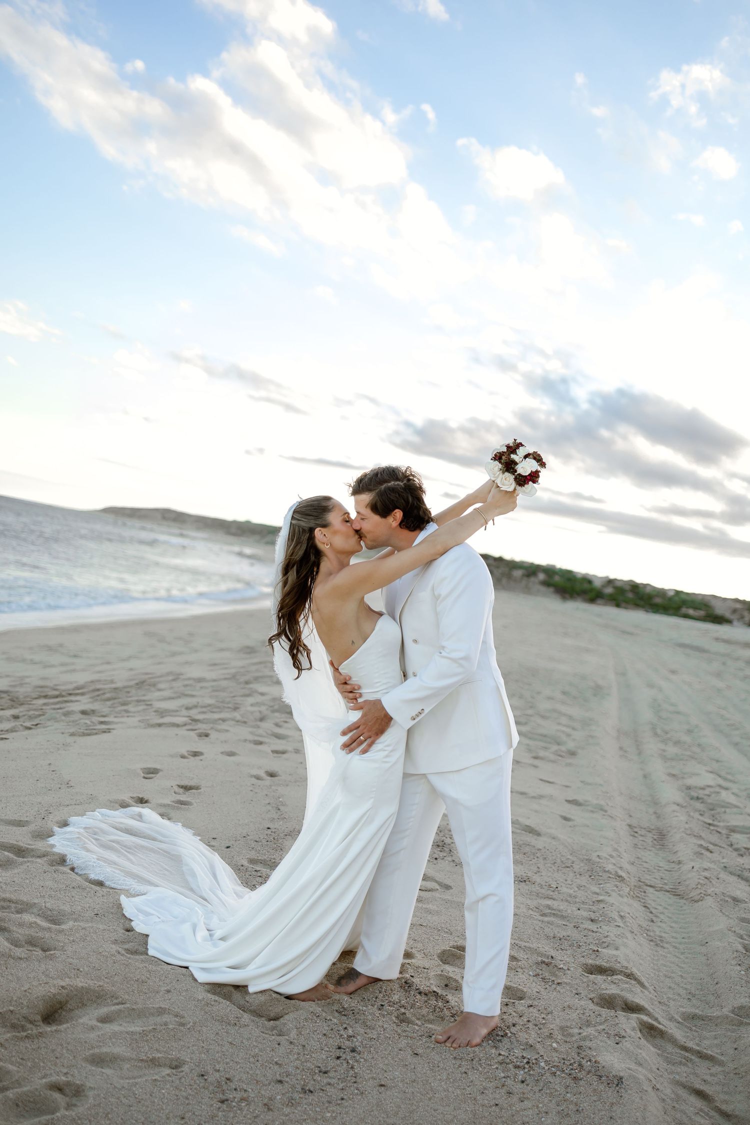 Bride and groom kissing on sandy beach at sunset, bride in white gown holding bouquet, groom in white suit.