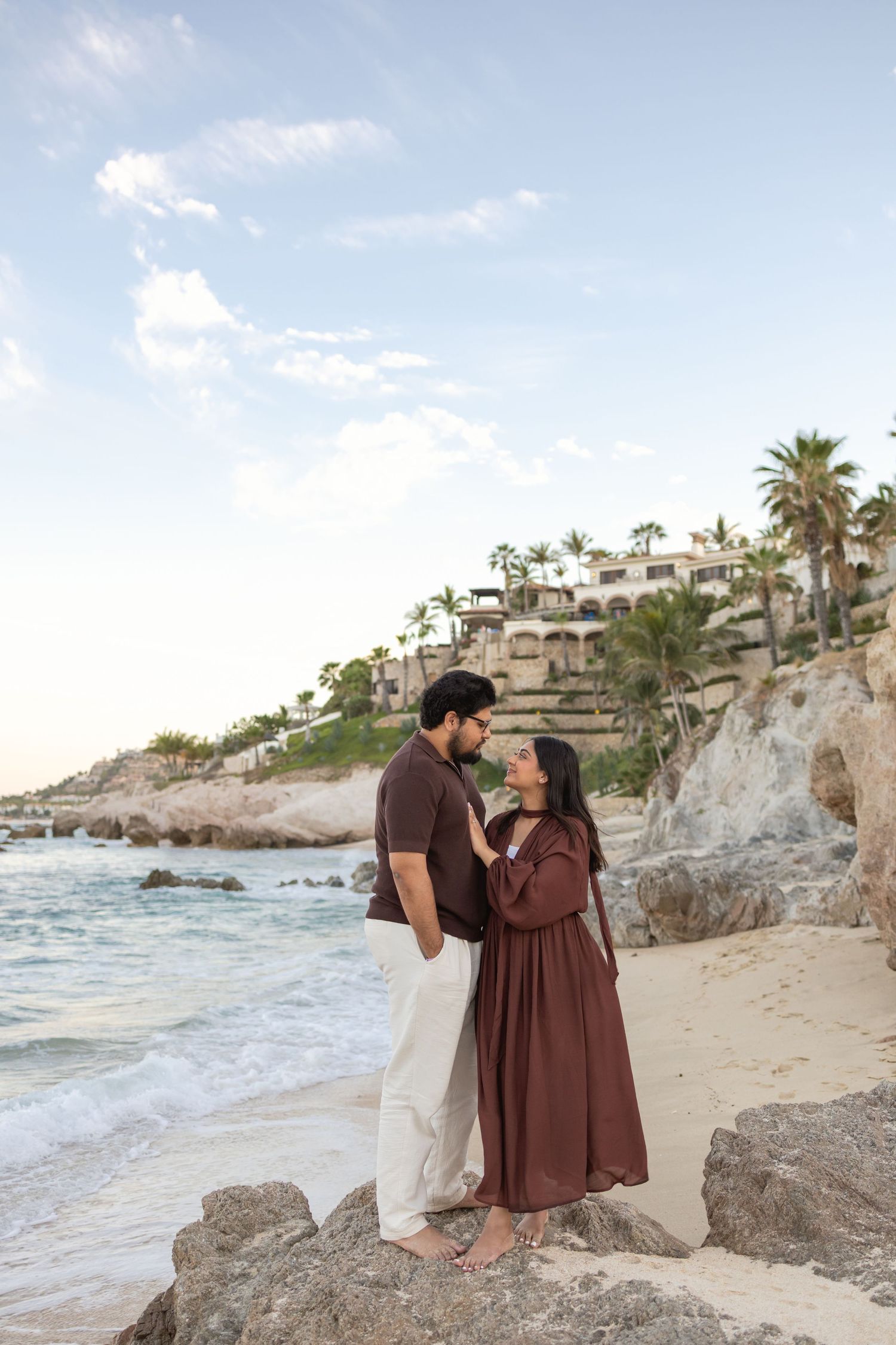 Couple in matching brown outfits share a romantic moment on a rocky beach with palm trees and resort in background.