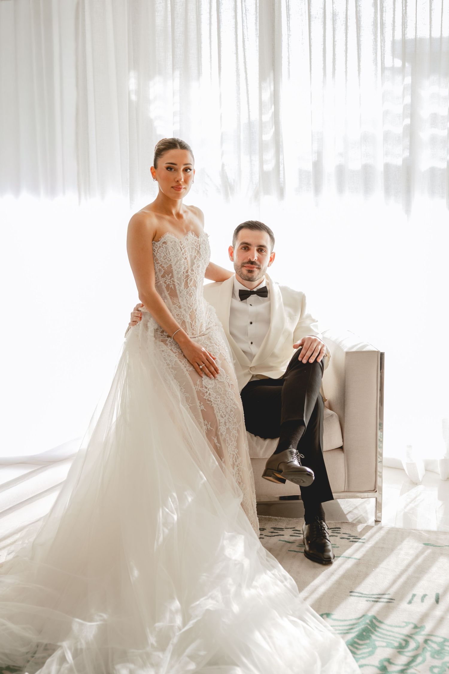 Bride and groom sitting together in sunlit room with flowing white dress.