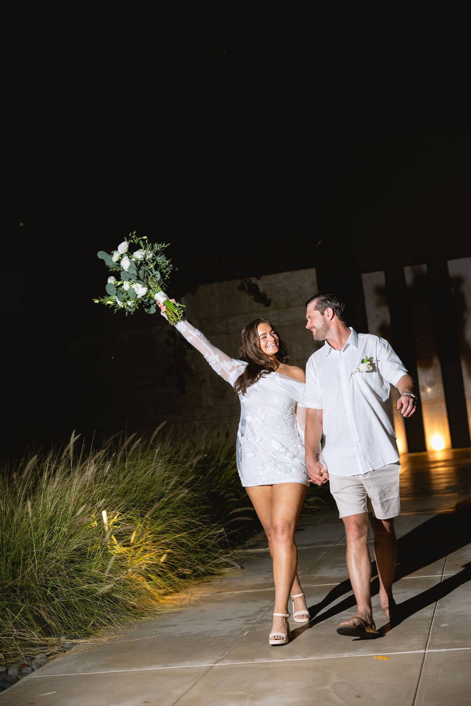 Couple in white outfits walking along path with decorative grasses at night.