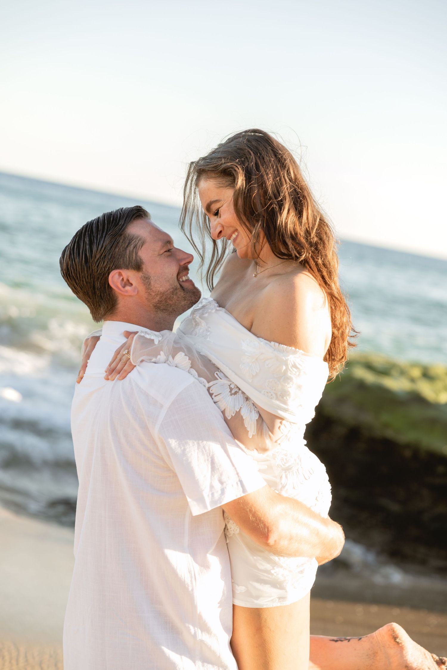 Couple in white sharing romantic moment during beach photoshoot at sunset.