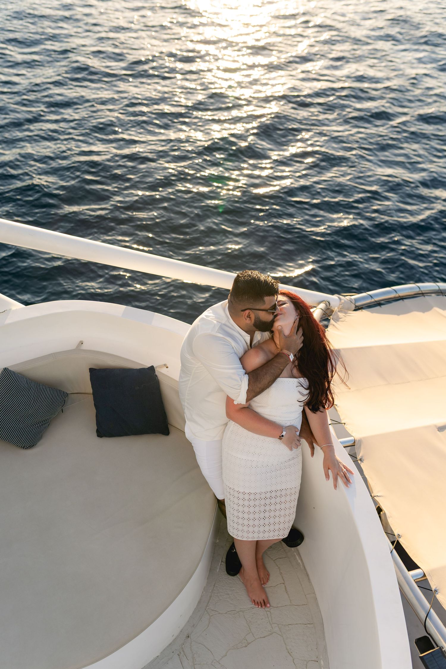 A romantic couple shares a tender moment on a white yacht deck with sparkling ocean waters in the background.