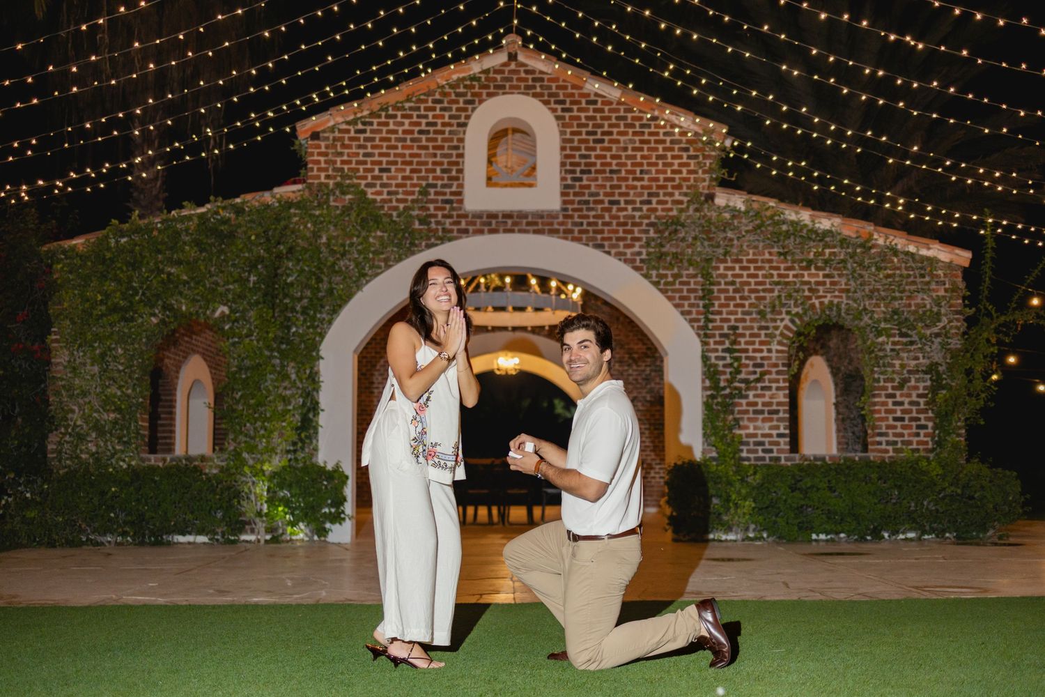 String lights illuminate a romantic outdoor wedding proposal scene at a rustic brick venue.