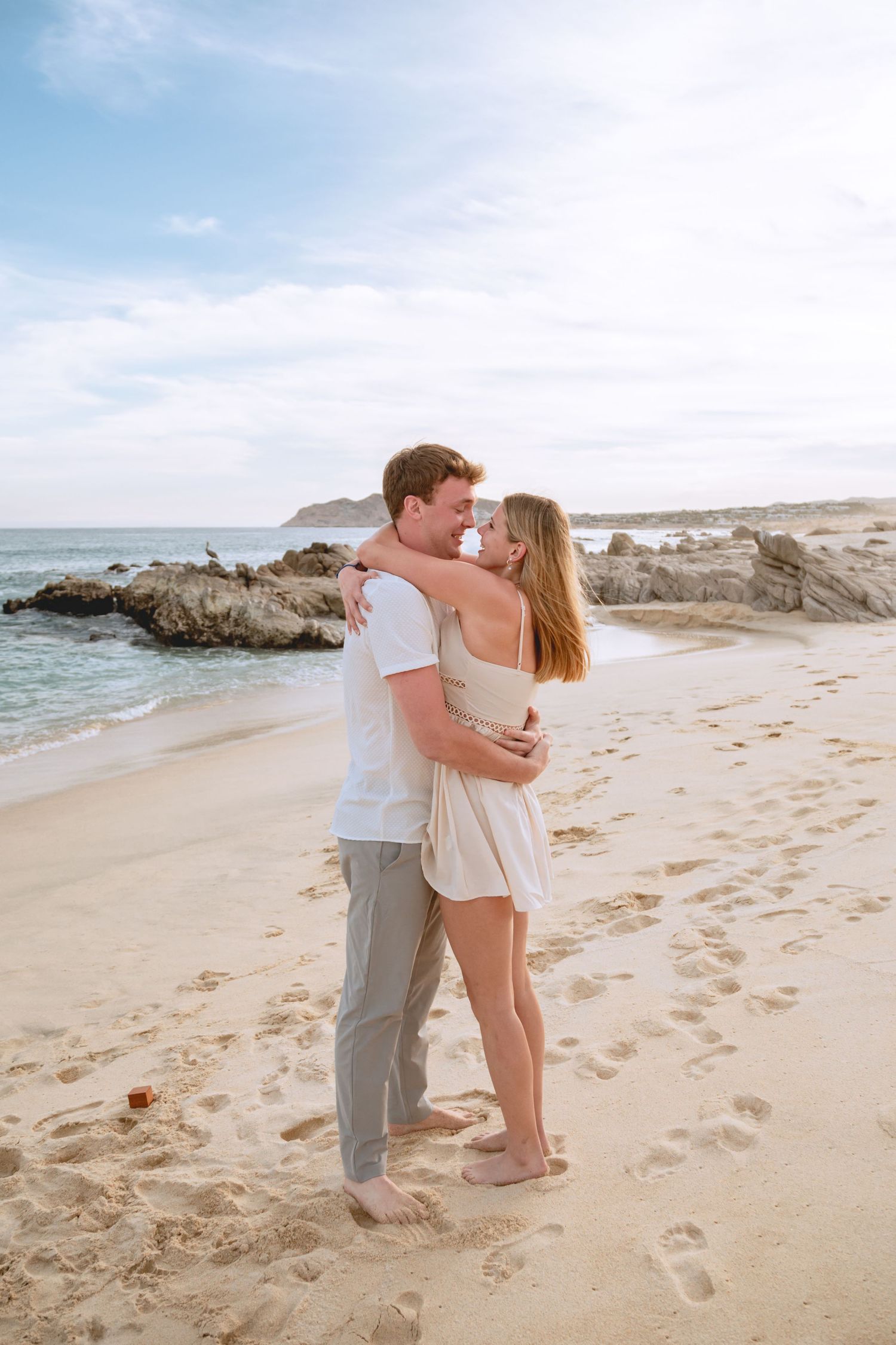 A romantic couple shares tender moments on a scenic beach with rocky outcrops and ocean waves in the background.
