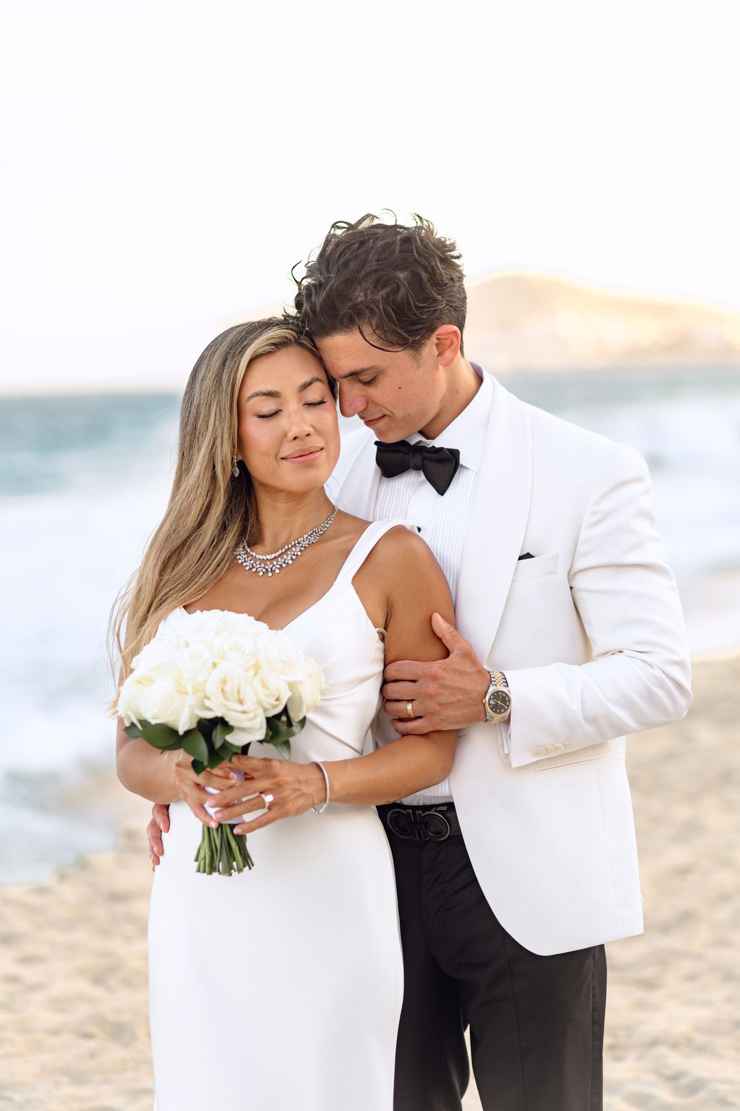 Intimate wedding portrait with couple holding white rose bouquet on beach.