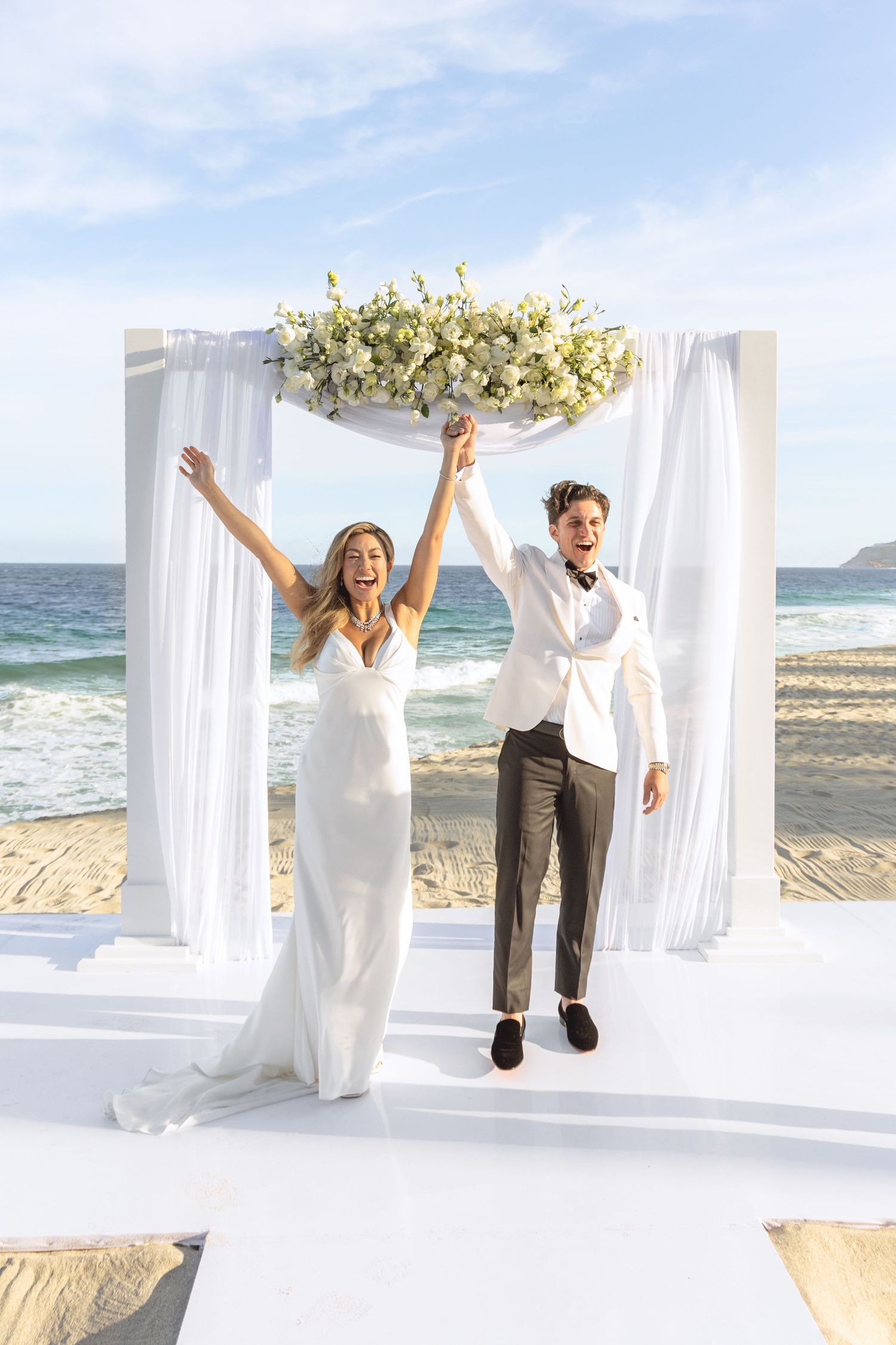Joyful couple celebrates under white beach wedding canopy with ocean backdrop.