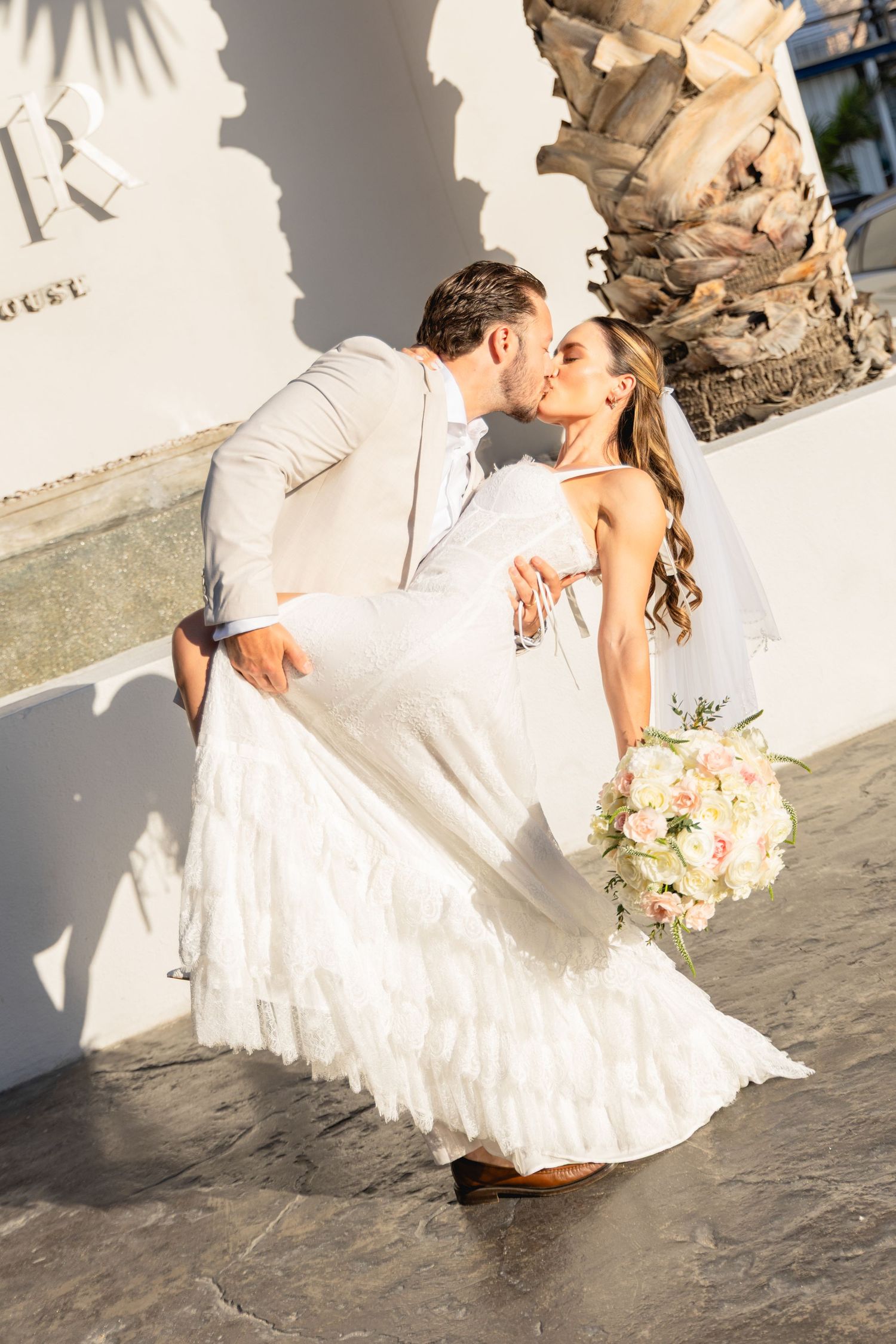 Newlyweds share romantic kiss while holding bouquet against white wall with palm tree shadows.