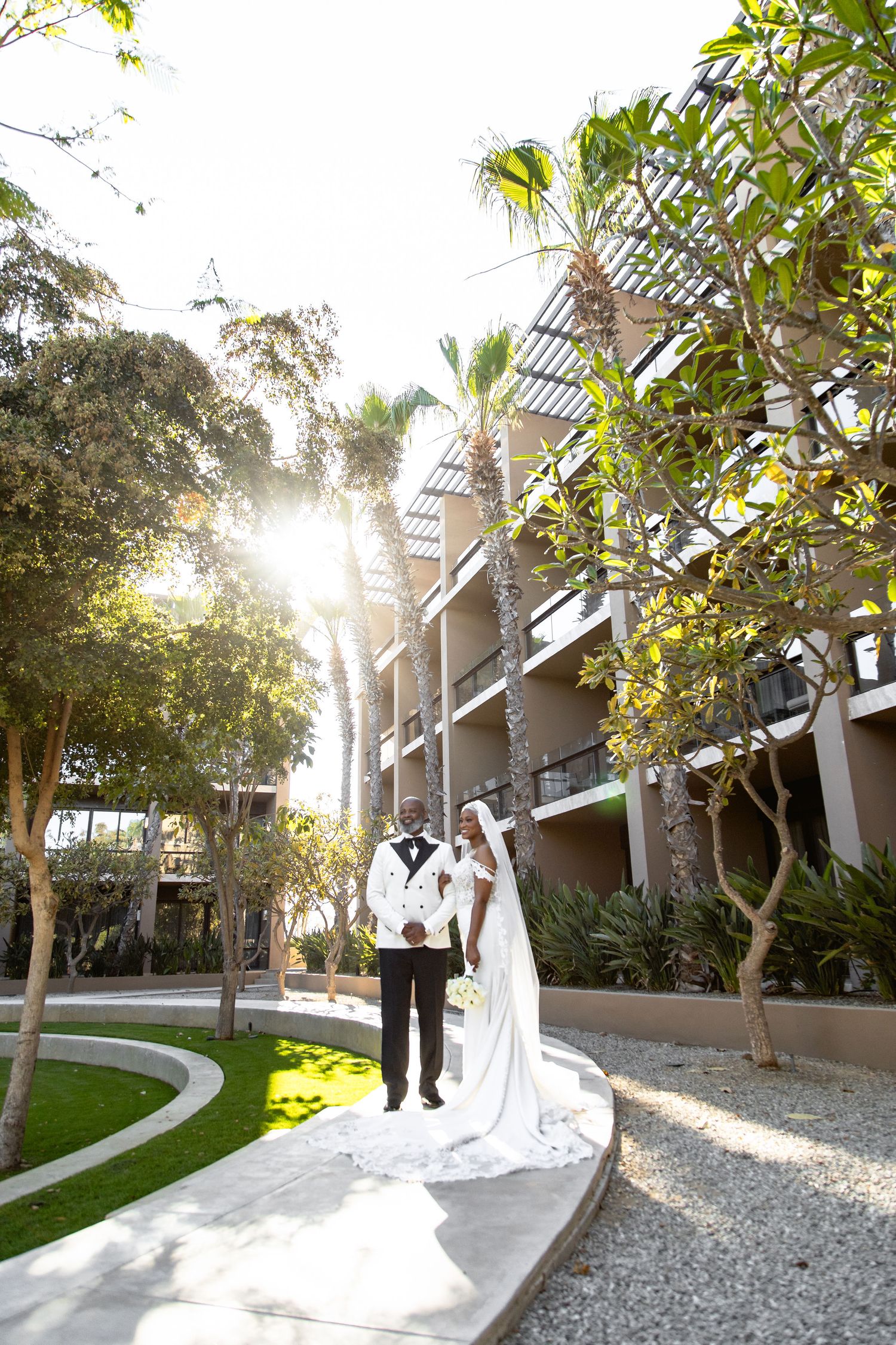 Newlyweds walking along garden path with sunlight streaming through palm trees.