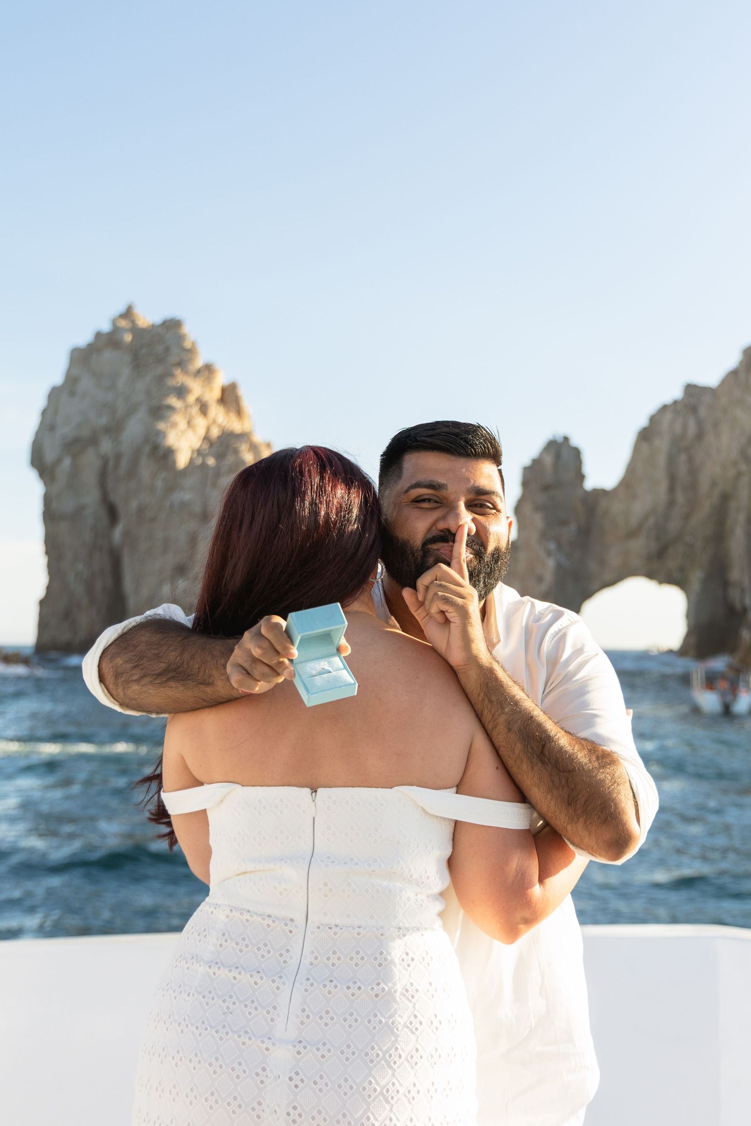 A couple shares an intimate moment against a backdrop of rocky ocean cliffs and blue water at sunset.