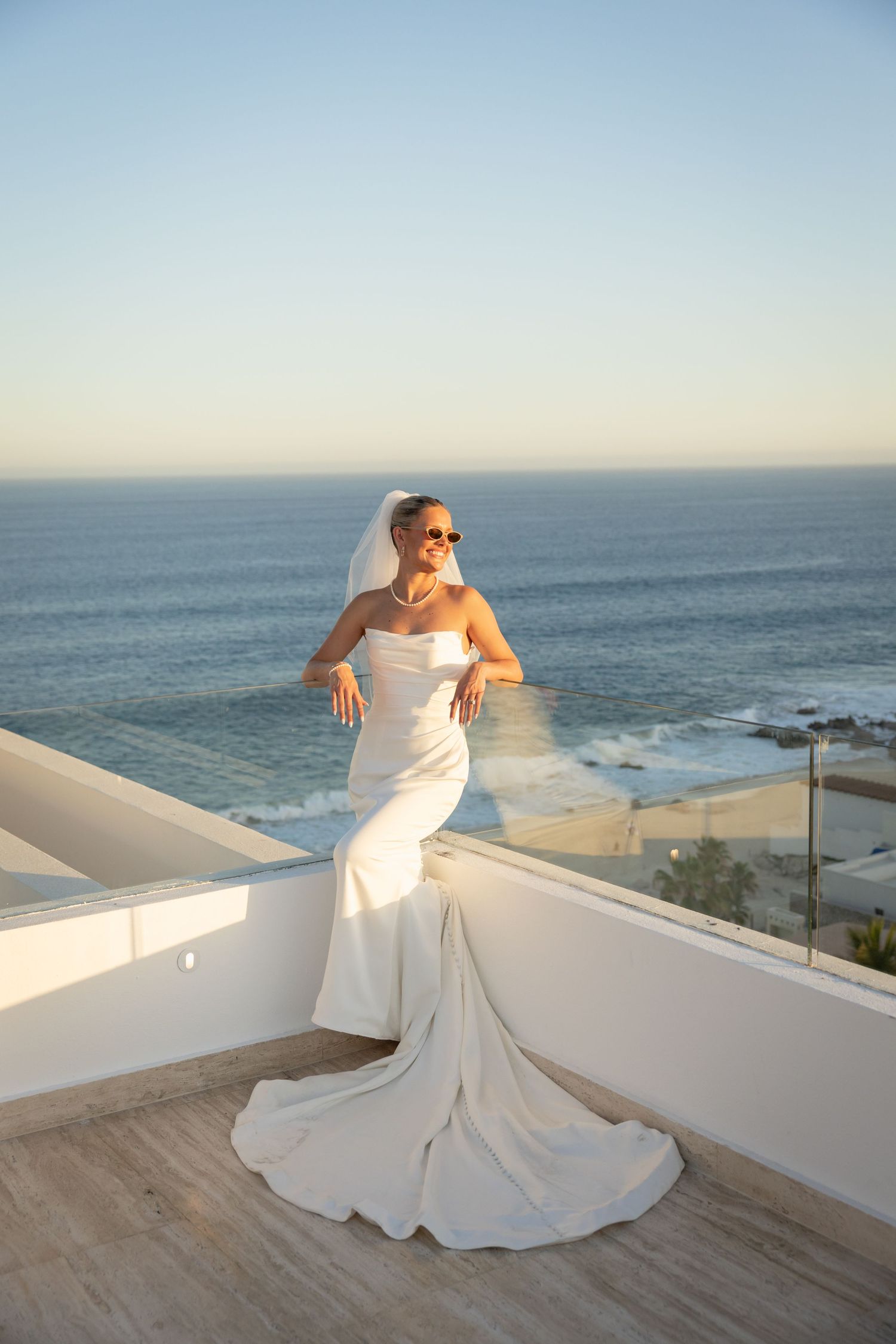 Bride in white strapless gown with veil and sunglasses poses on rooftop terrace overlooking ocean at sunset.