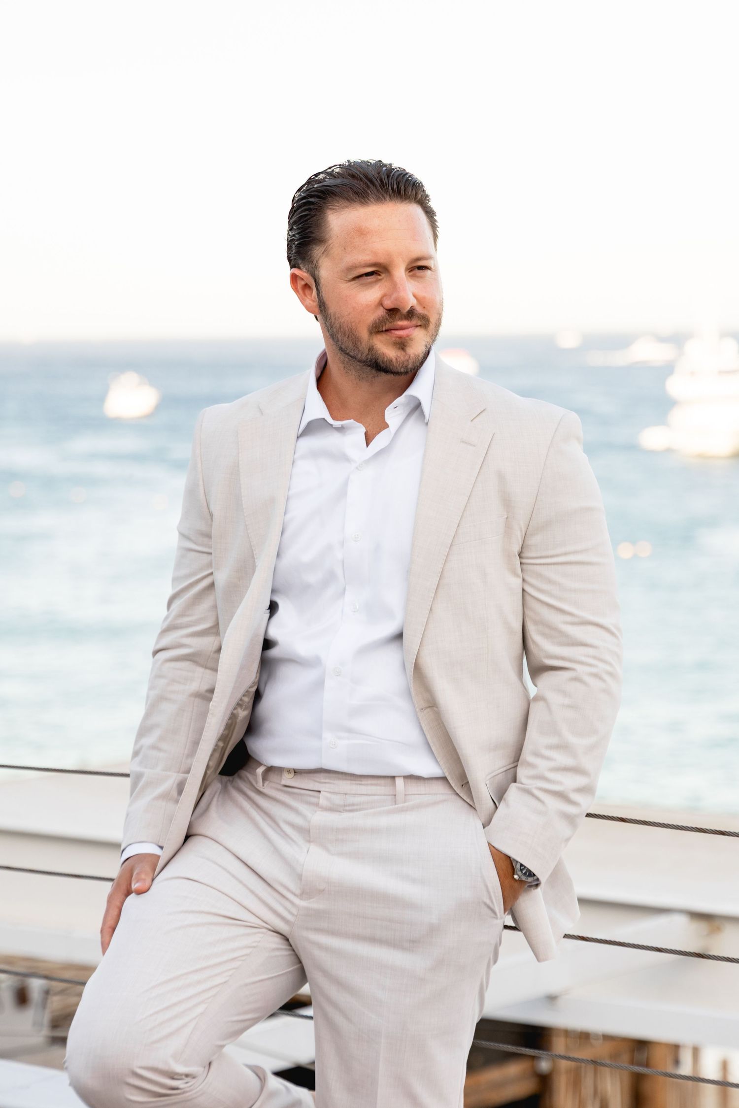 Elegant groom in light beige suit poses by the ocean.