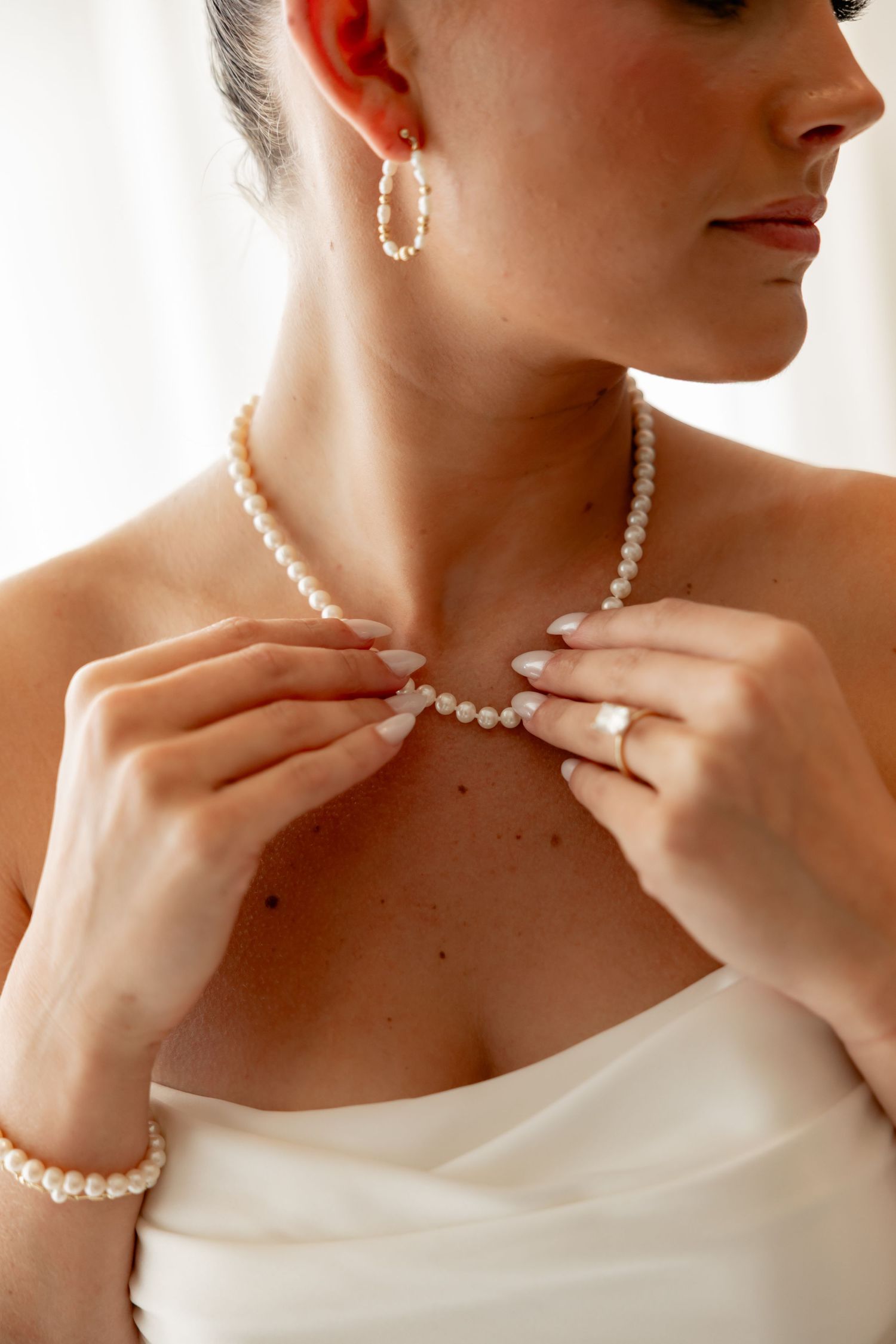 Bride adjusting pearl necklace wearing strapless white dress, pearl earrings, bracelet, and diamond ring.