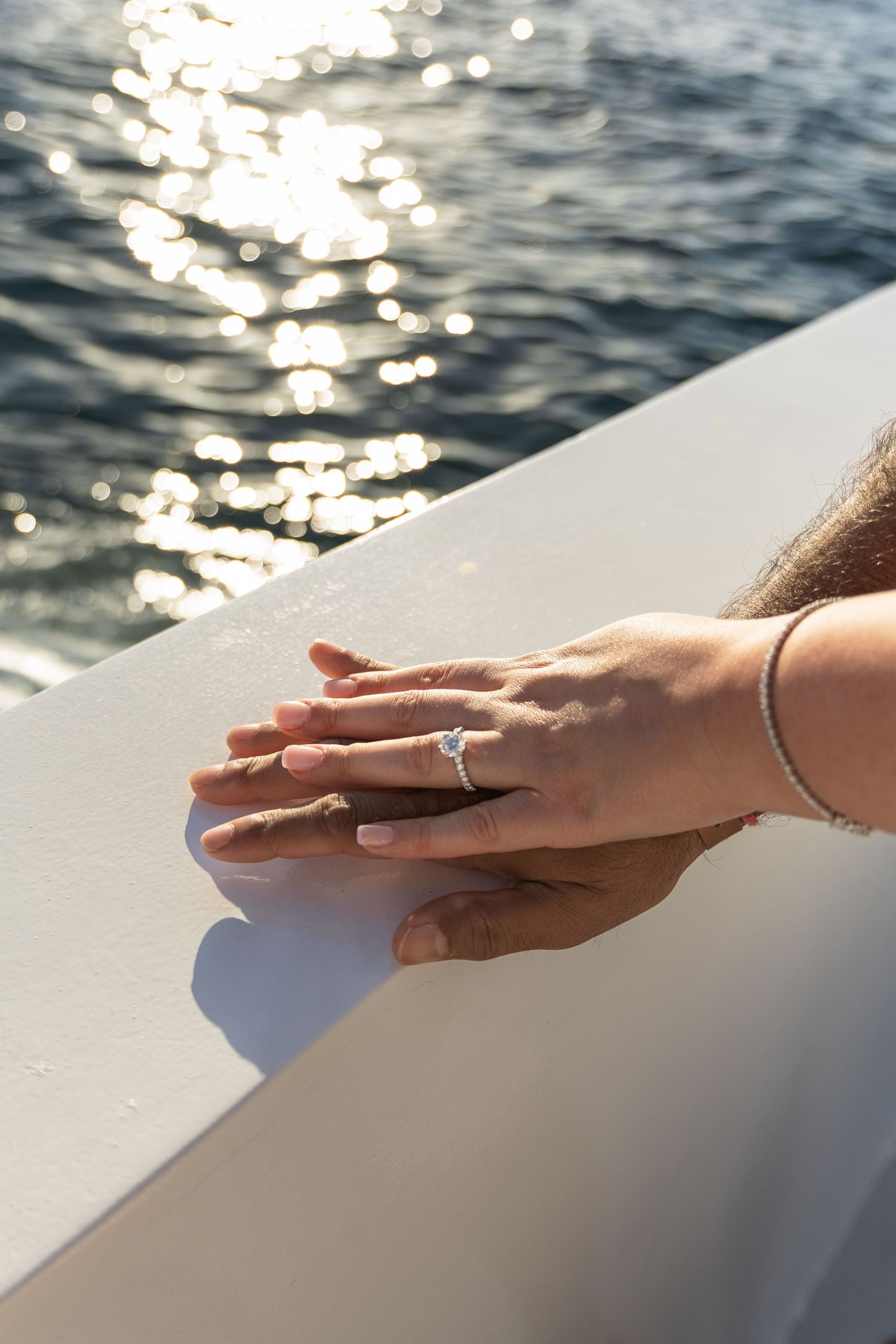 Hand with engagement ring and bracelet rests on white surface near sparkling water in sunlight.