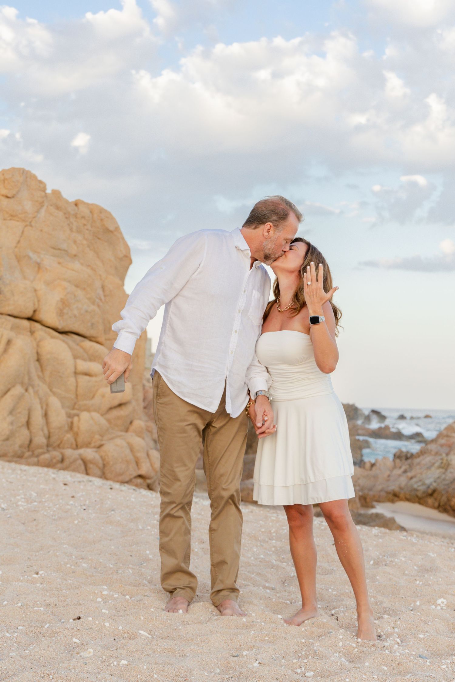 A couple in white and khaki outfits shares a romantic moment while walking along a sandy beach with rocky cliffs.