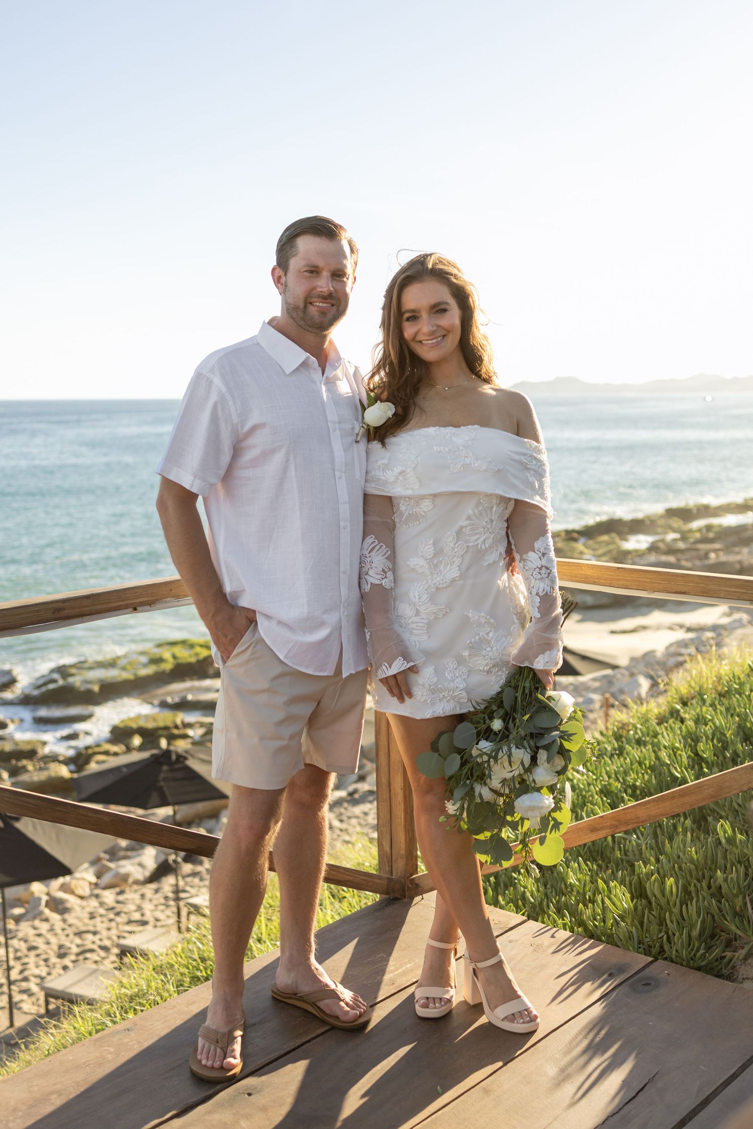 A couple in white beachwear poses together on a coastal boardwalk with ocean views.