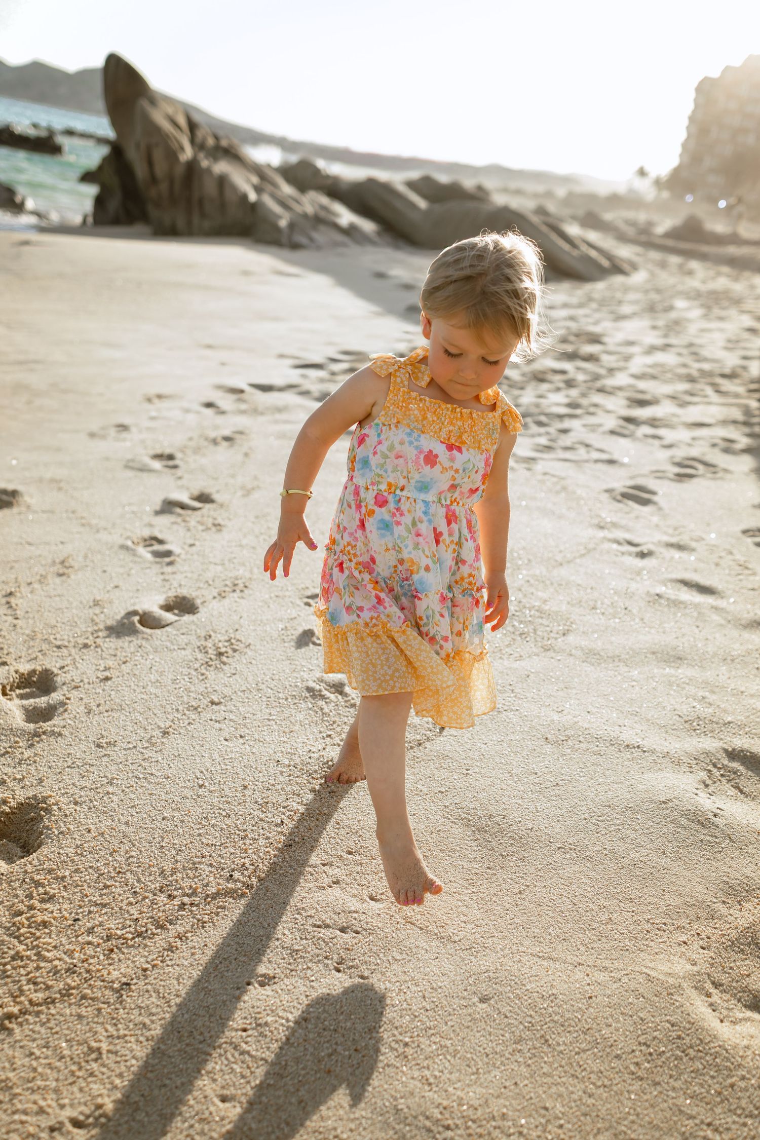 Young child in floral sundress walks barefoot on sandy beach during golden hour, casting long shadow at Garza Blanca, Los Cabos