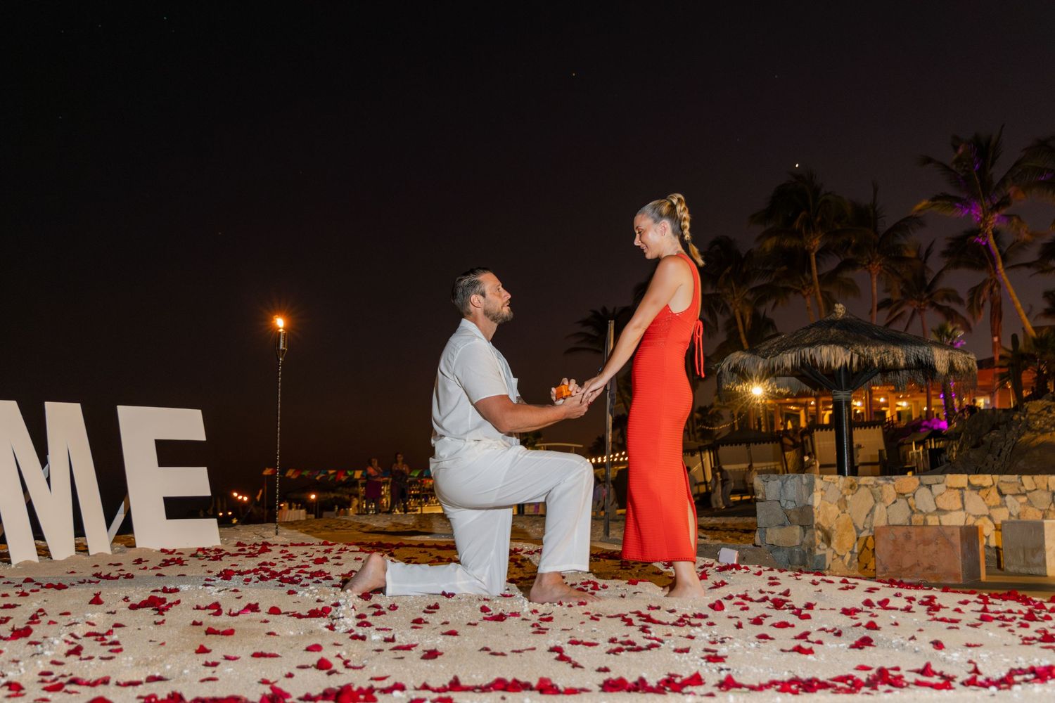 A romantic proposal scene at night with rose petals scattered on the ground near illuminated ME letters.