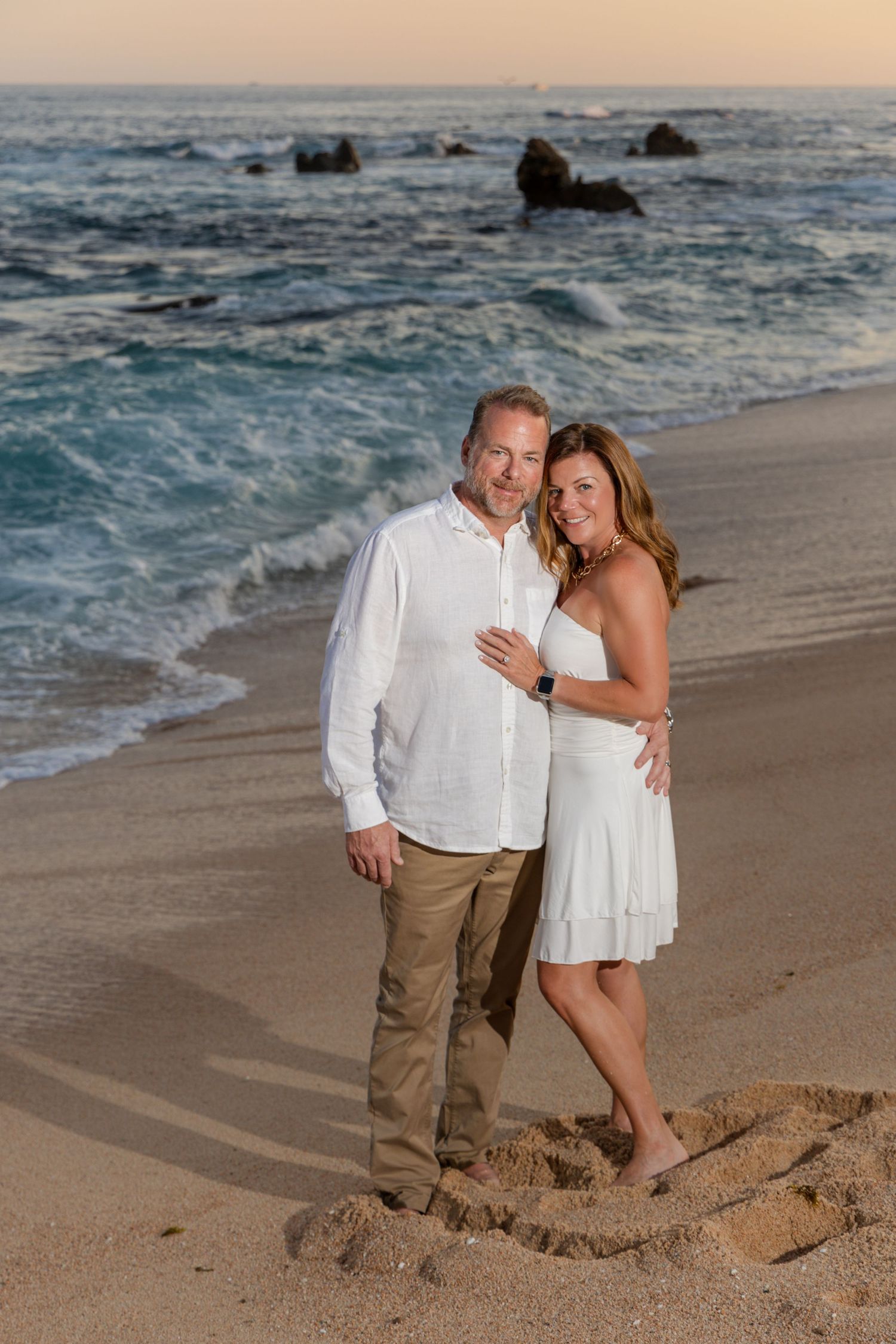 A couple in white attire poses romantically on a sandy beach at sunset with ocean waves in the background.