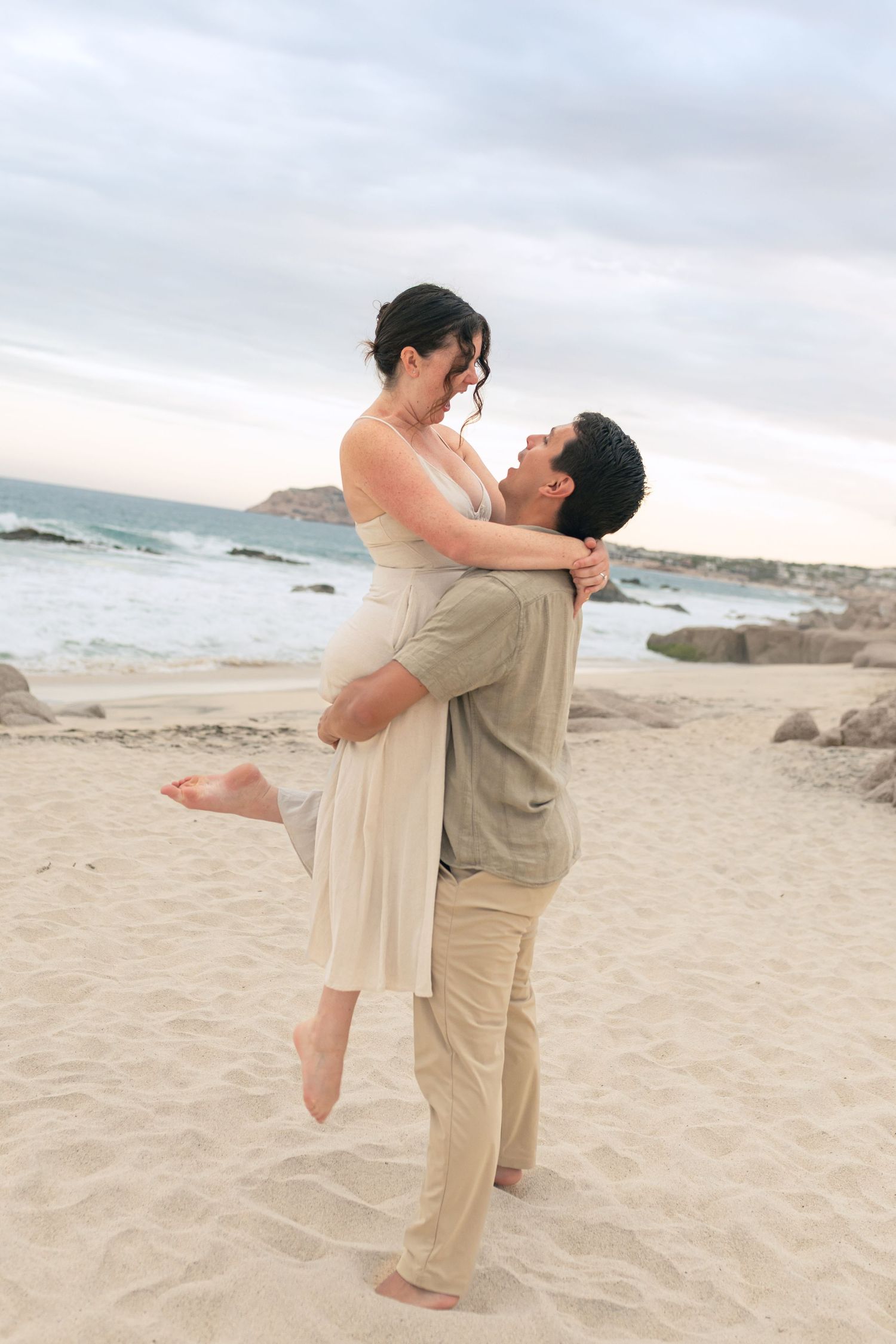 A romantic couple embraces and spins joyfully on a sandy beach at sunset with ocean waves in the background.