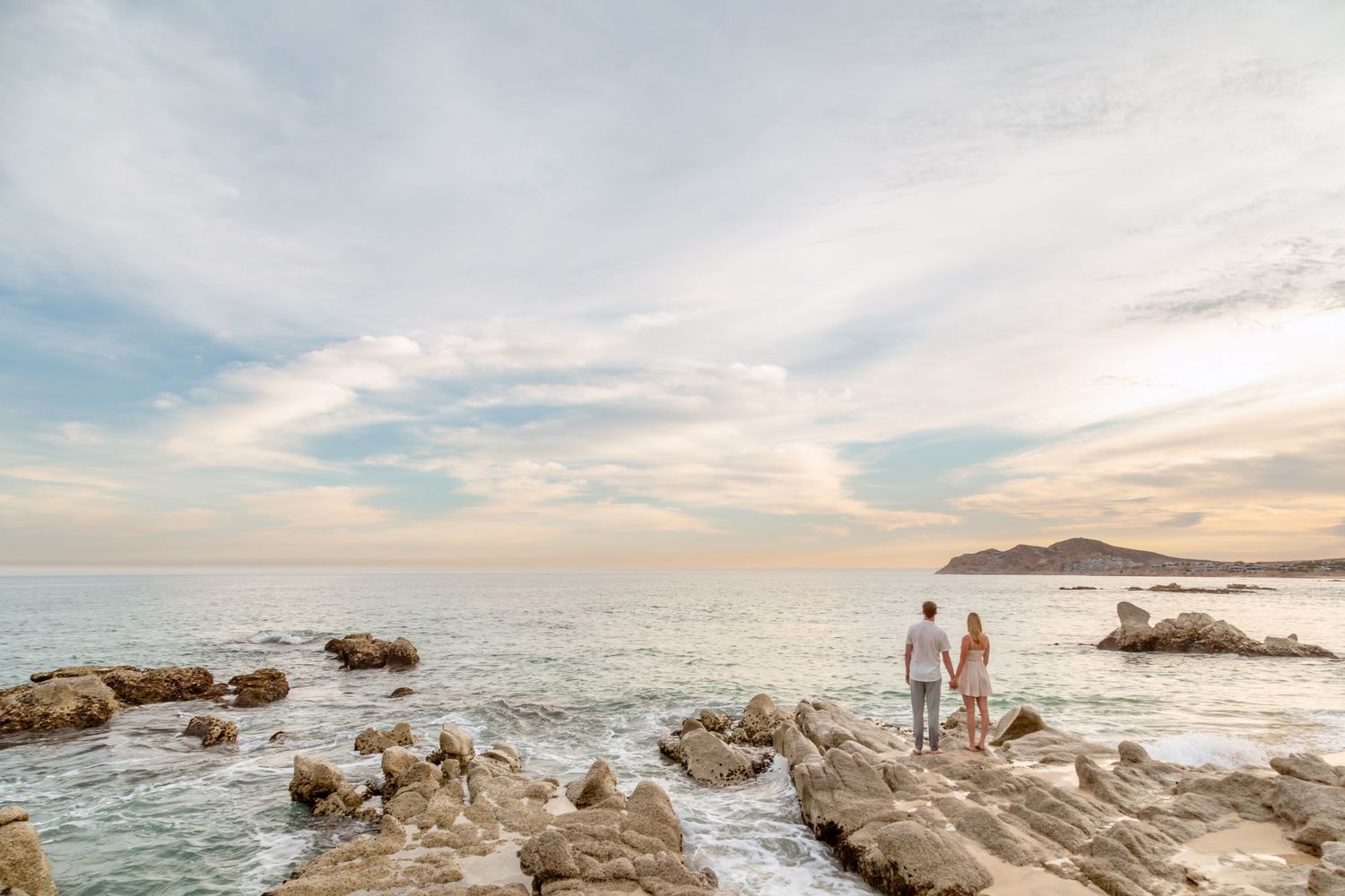 Two people stand on rocky coastal outcrop overlooking ocean waves and distant mountains at sunset.