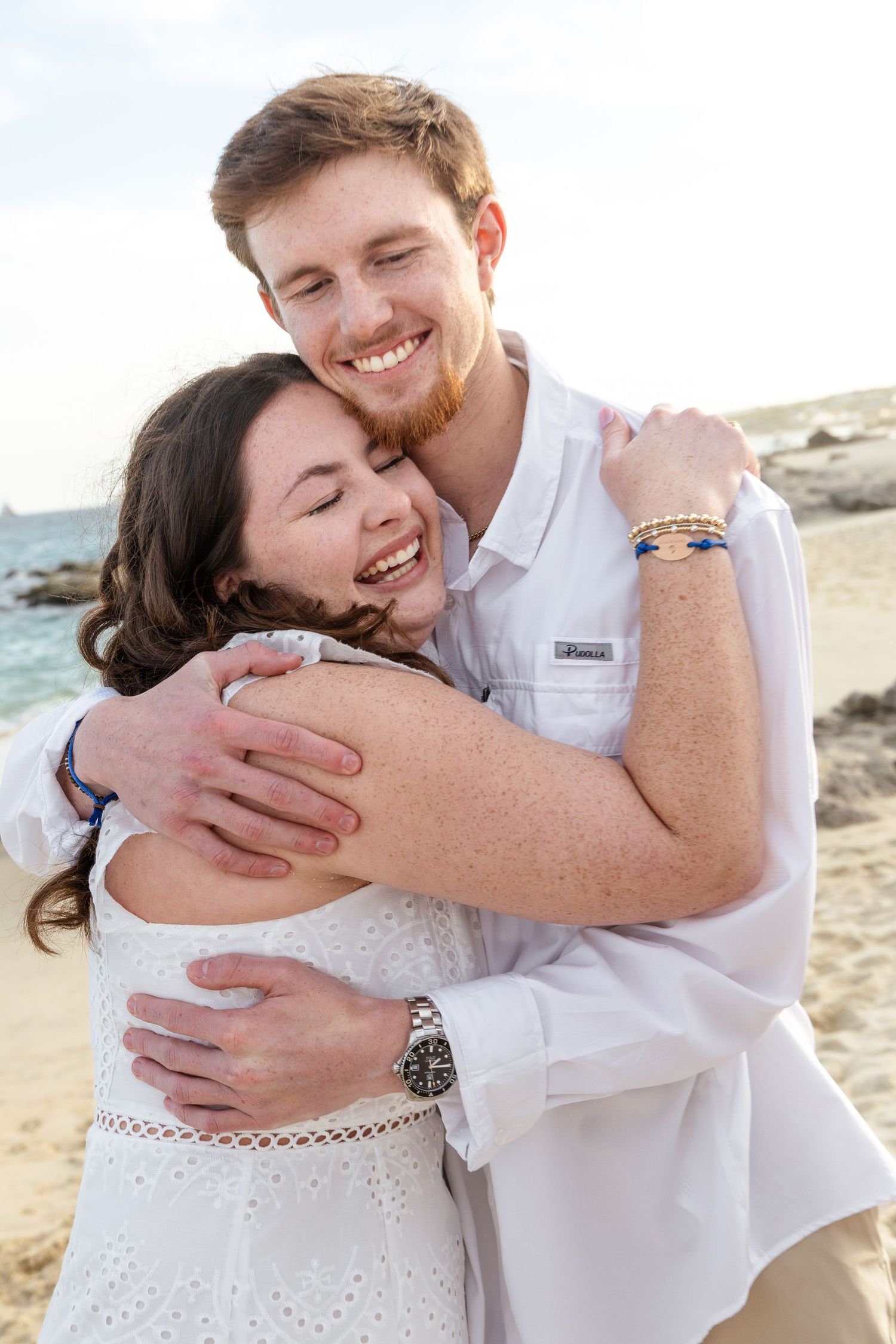 A couple embraces and laughs together during a beach photoshoot in white attire.