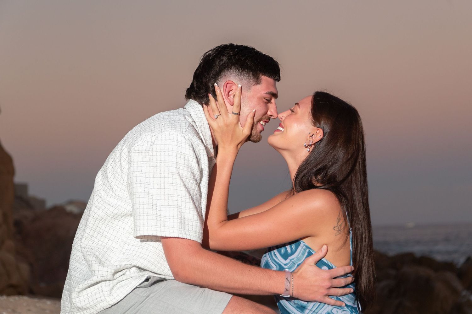 A romantic couple shares tender kisses against a sunset backdrop at the beach.